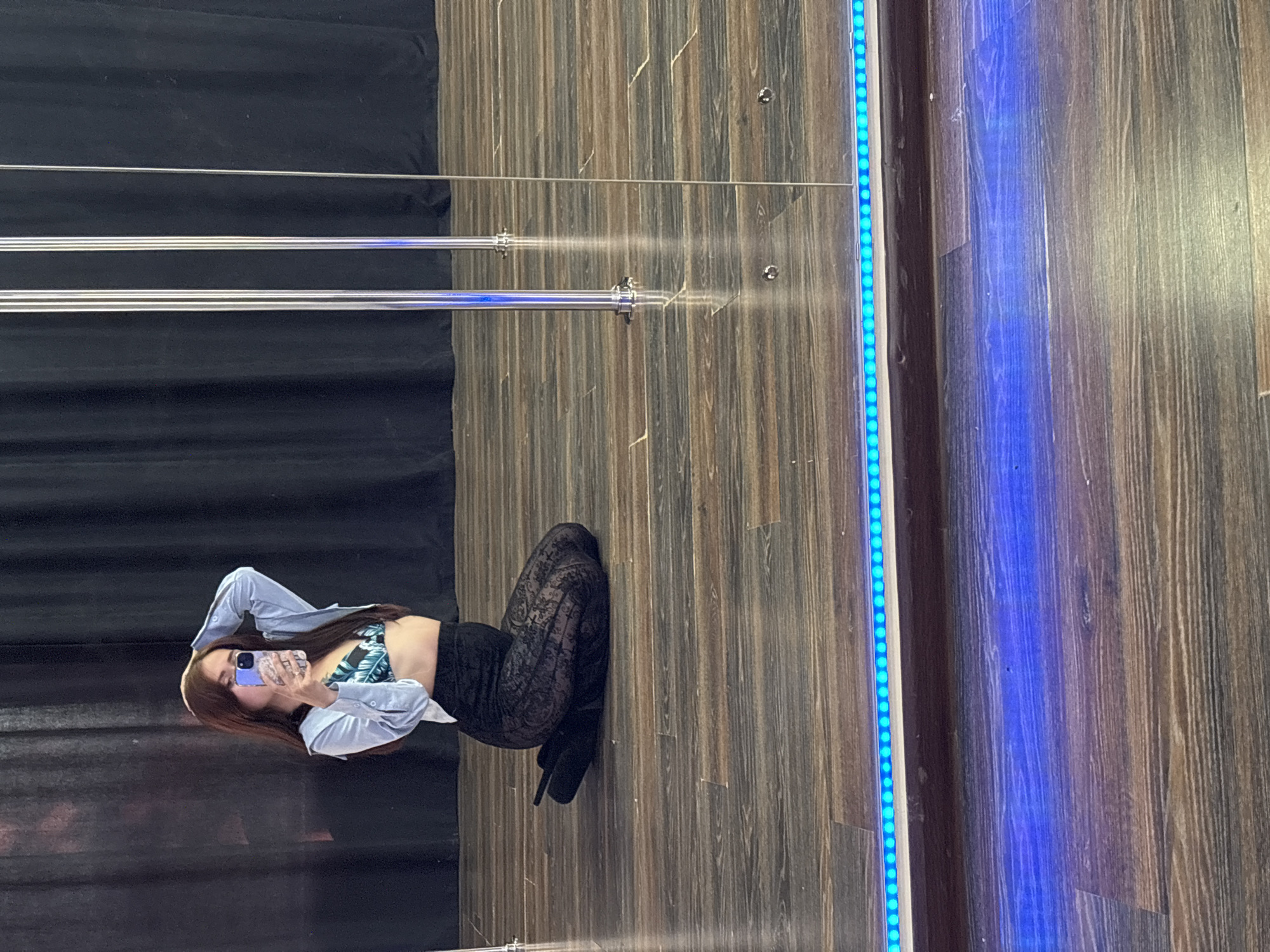 A woman is lying on the floor, taking a selfie with her phone. She is wearing a light blue shirt, a bikini top with a leaf pattern, and black lace pants. The scene appears to be indoors, with a dark curtain in the background and a wooden floor. To the right of the woman, there is a wall with blue LED strip lights. The image is taken in Naberezhnye Chelny, Russia. The lighting suggests it could be daytime or artificial lighting. The woman's pose is relaxed and informal. There is no visible text in the image.