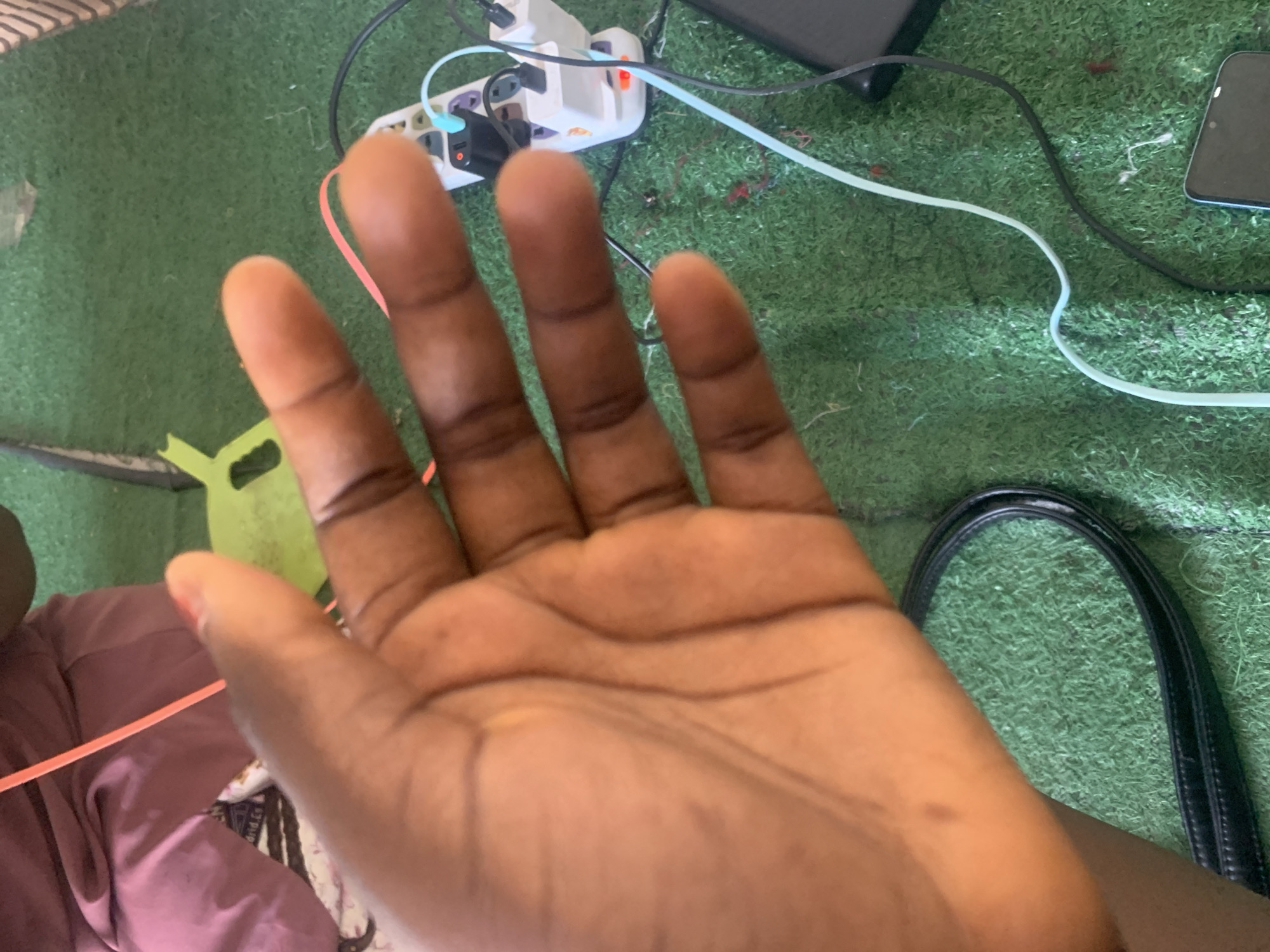 In Jauro Yinu, Nigeria, a close-up, overhead shot captures a person's right hand, palm facing upward and fingers slightly curled. The hand is dark-skinned and shows prominent lines on the palm. In the background, artificial green turf covers the ground. A white power strip with multiple sockets is visible, plugged into which are various cables and adapters, including a black device and a red indicator light. A pink cord snakes across the scene. To the left, a person's legs are clad in purple pants. A black strap, possibly from a bag, lies on the turf near the bottom left of the frame. The image is focused on the hand, with the background elements slightly blurred.