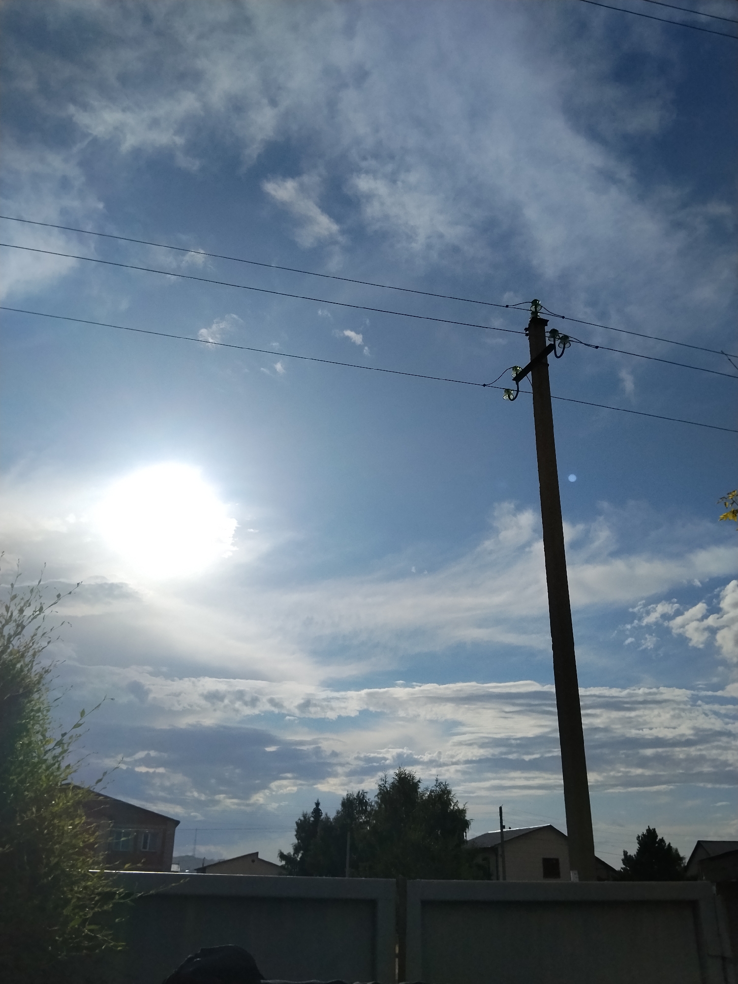 The image captures a bright, sunny day with a vast expanse of blue sky filled with wispy white clouds. The sun is prominently featured on the left side of the frame, emitting a strong, bright light. In the foreground, a power pole stands tall, supporting electrical wires that stretch horizontally across the sky. The pole is made of concrete and has insulators attached to it. Below the sky and power pole, a residential area is visible, with houses and trees. A fence runs along the bottom of the image, partially obscuring the view of the houses. On the far left, a bushy green plant is in sharp focus. The overall scene suggests an outdoor environment in a town or city. This image is located in Kokshetau, Kazakhstan, specifically in the southwestern part of the city, near the lake Shchuchye.