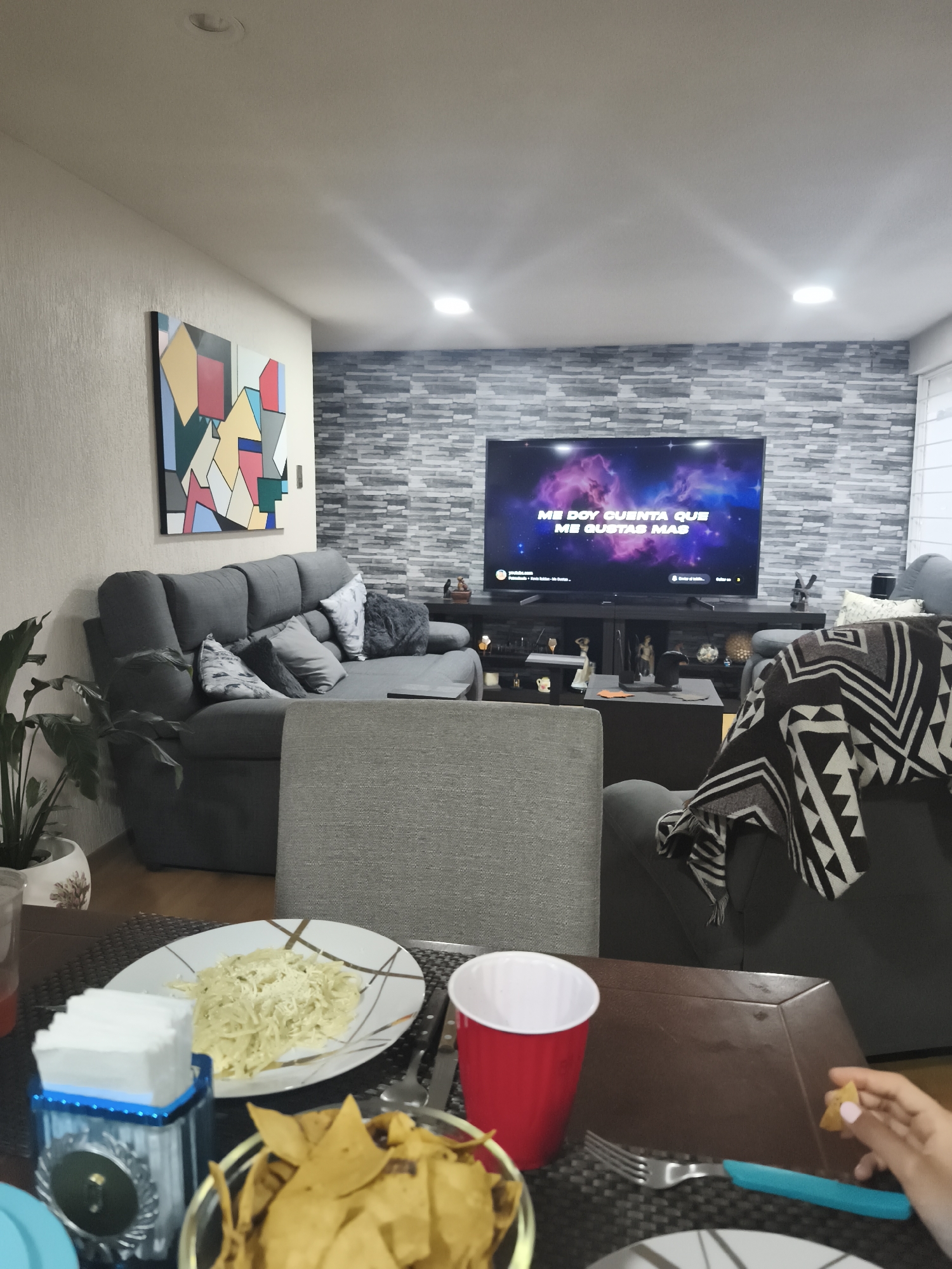 The photograph shows an interior scene likely taken in a living or dining area of a residence. The room is furnished with a gray sectional sofa, a dark entertainment center, and a dining table. A gray chair is also visible. The walls feature a textured gray brick design behind the TV and a contrasting off-white paint. There is abstract artwork on the wall to the left.
The television displays the phrase "ME DOY CUENTA QUE ME GUSTAS MAS." On the dining table, there is a plate of pasta, a bowl of tortilla chips, a red cup, and a blue napkin holder. A person is holding a tortilla chip.
Given the phrase on the TV screen is in Spanish, and the fact that the chips are a staple of Mexican cuisine, it is possible that this image was taken in Mexico. However, it is not possible to determine with certainty whether the image was taken in Mexico City without more specific information. The presence of tortilla chips suggests it's possible that this image was taken in Mexico City, Mexico.