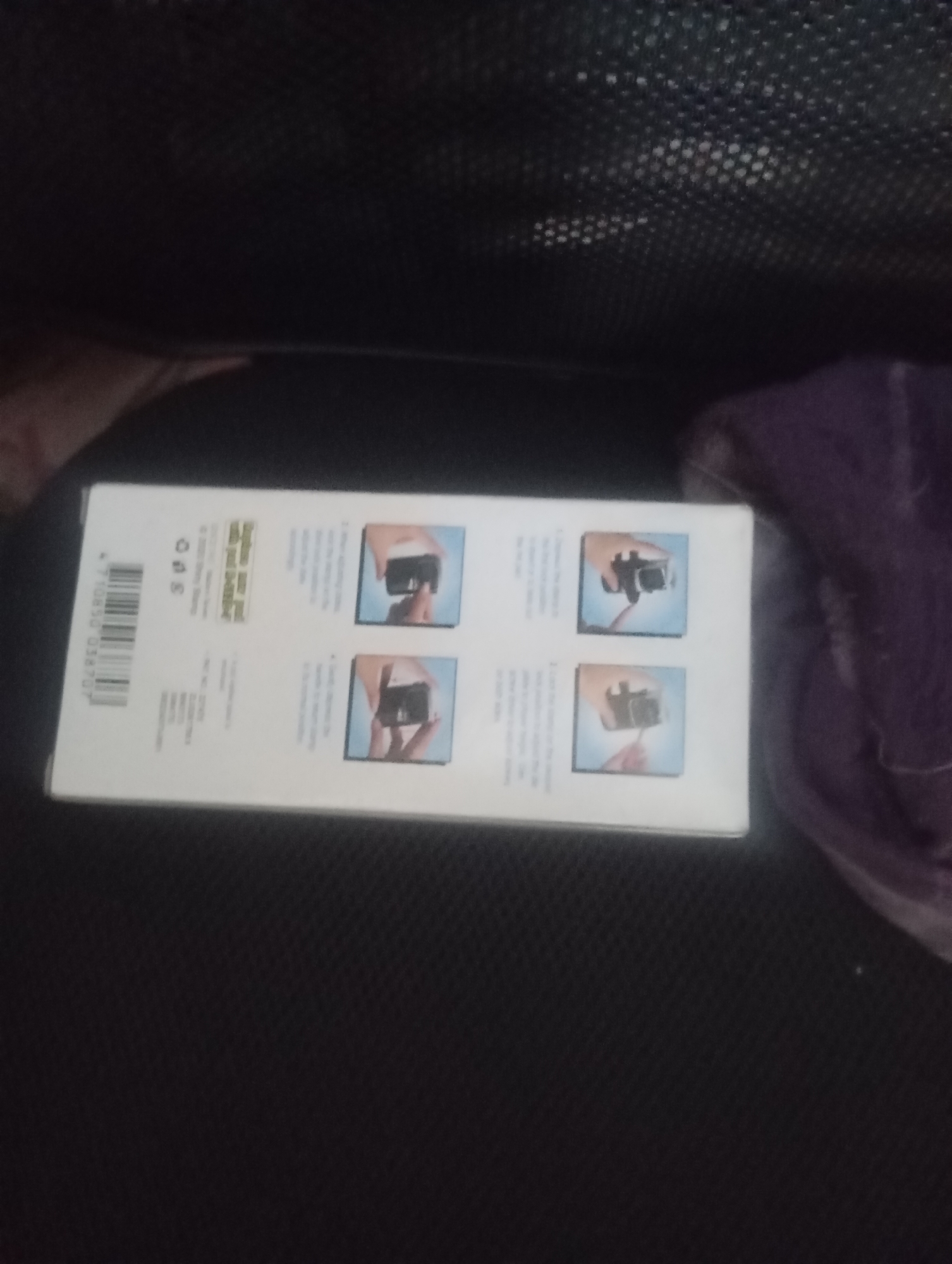 The image shows a close-up of a white box with product information and four small instructional images. The box has text and a barcode on the left side, and on the right side, there are four square images depicting hands holding and using a small black device. The overall image is slightly blurry and taken from an angle, with the box resting on a dark, textured surface. The background is dark and out of focus. There are no discernible features that would allow for the identification of the city of Jos, Nigeria, or any specific environmental details related to it.