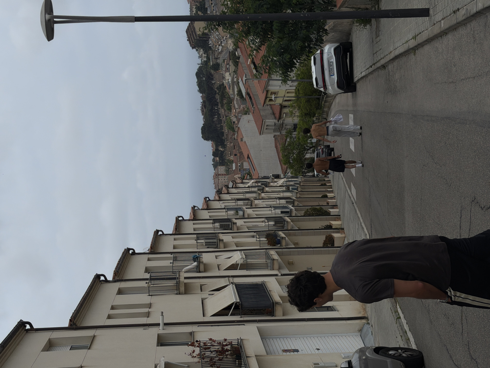 The image shows a street in Sant Andreu de Llavaneres, Spain. The view is from a high angle, looking down a steep street with apartment buildings on the left side. The buildings are white with multiple balconies. On the right side of the street, there's a row of trees and older buildings, with red-tiled roofs.
A light post stands at the top left of the image. In the foreground, a person wearing a dark gray shirt and black track pants stands looking down the street. Further down the street, two shirtless men are jogging away from the viewer, one in gray pants and the other in black shorts. A white car with license plate "2086 NFN" is parked along the right side of the street, near the running men. Another car can be seen at the base of the image.
The sky is overcast. The overall impression is that of a residential street in a hilly area.