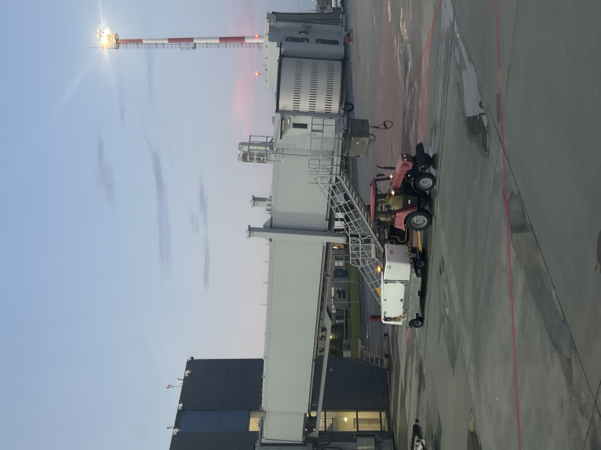 This media file depicts an airport scene at dusk, with a clear sky and the early stages of twilight visible. The main subject is an airport jet bridge, a long, elevated walkway that connects an airport terminal to an aircraft. The jet bridge is a light gray color with white sections and metal stairs leading up to it.
Adjacent to the jet bridge is a tow tractor, a small, red vehicle used for moving aircraft and ground support equipment. The tow tractor appears to be connected to a white piece of equipment, possibly a baggage or cargo loader, which has the text "TID" and "655" visible on its side. A person in a yellow vest is visible near the tow tractor, likely an airport worker.
In the background, a tall tower with red and white stripes stands prominently, illuminated by a bright light at its peak, suggesting it's a navigation aid or communication tower. Other airport structures, including a dark-colored building with windows, are also visible. The ground is paved and wet, indicating recent rain or damp conditions. The overall atmosphere suggests a quiet moment at the airport before the evening rush or after a flight has departed. No specific emotions are conveyed, and the scene is factual and functional.
