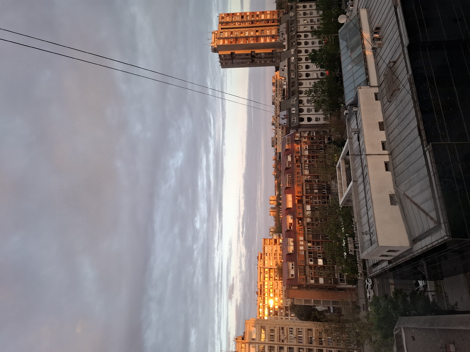 The image depicts a cityscape, likely taken at sunset, as evidenced by the warm light illuminating the buildings. The location appears to be Novi Sad, Serbia, judging by the architectural style of the buildings. Several high-rise residential buildings are visible, with numerous windows reflecting the setting sun. A utility line is strung across the top of the frame, silhouetted against the cloudy sky. In the lower right corner, a rooftop or adjacent building is visible, with visible equipment.