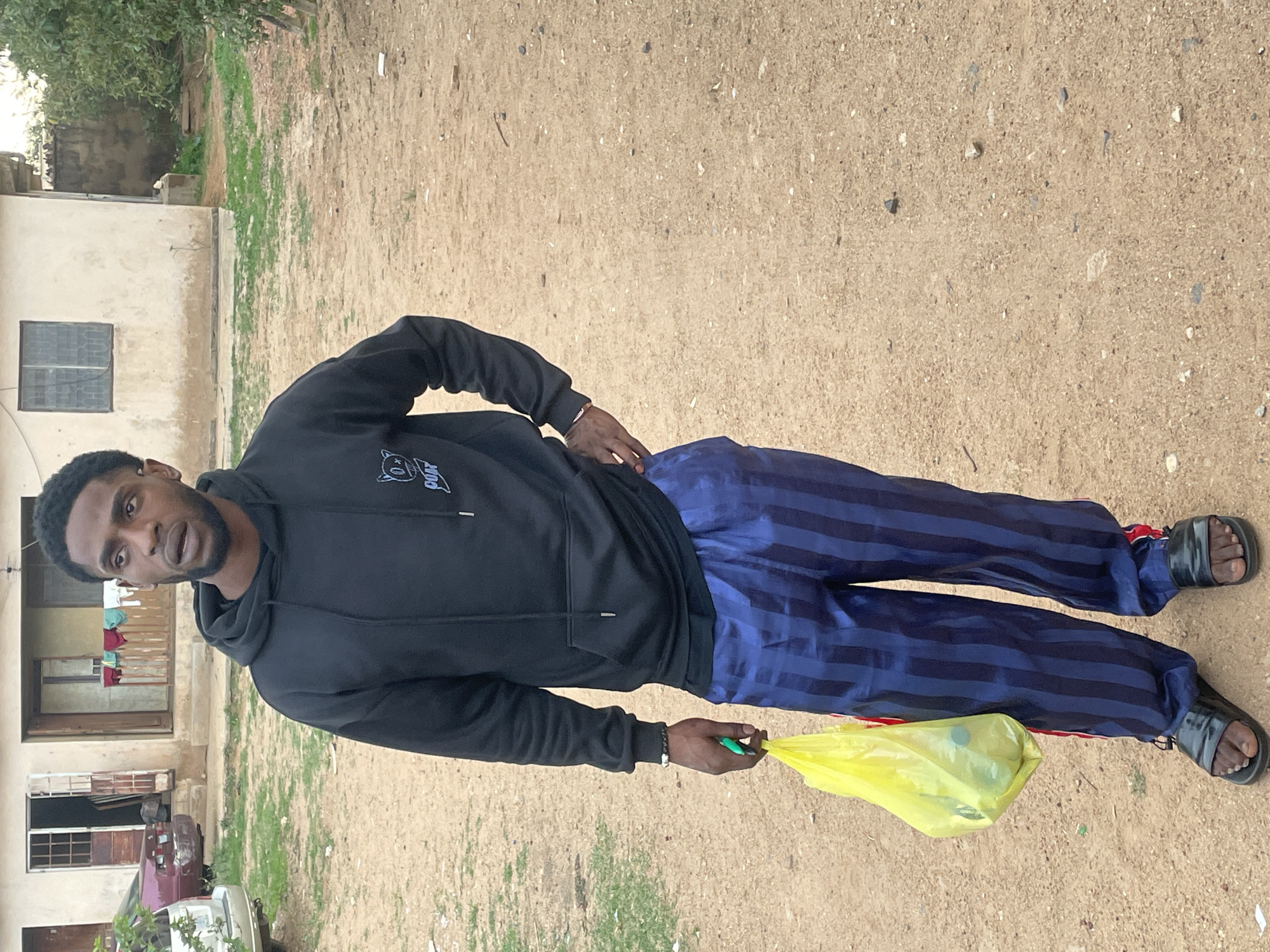 A man is standing outdoors in Jos, Nigeria. He is wearing a black hooded sweatshirt, blue striped pants, and sandals. He is holding a yellow plastic bag in his right hand. The ground is dirt and gravel. In the background, there is a building with windows and a red car parked on the side of the building. The environment appears to be a residential area.