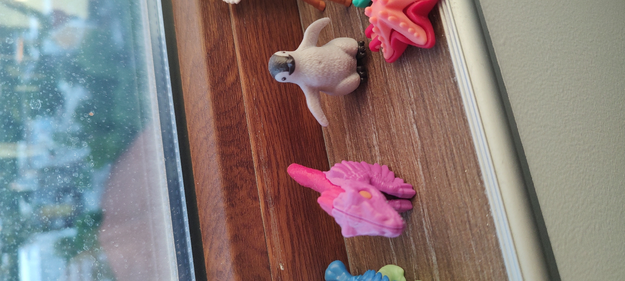 The image shows a window sill with several small toys. The window itself is slightly dirty, with a blurred cityscape visible through it. The toys include a white animal figure with black markings, a pink starfish, and a pink dinosaur. The window sill is made of brown wood and has a white frame. The toys are arranged haphazardly on the sill. The background wall is a light gray color. The location is Unknown, Unknown.