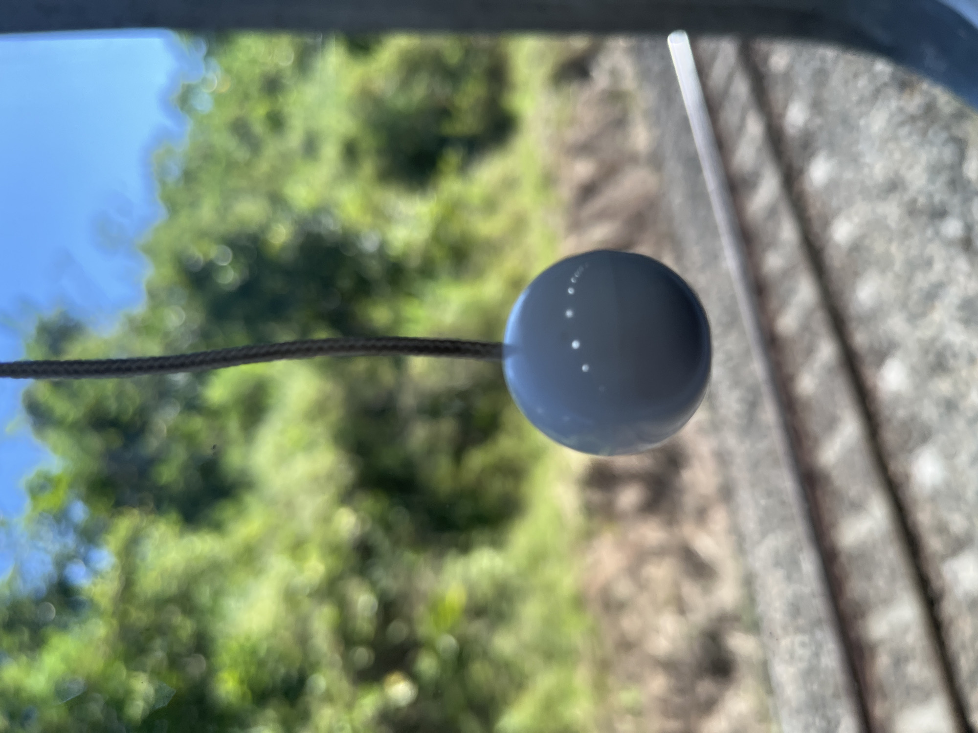A grey, spherical object, likely a plumb bob, hangs from a dark string in the foreground, suspended in front of a blurred background of lush green foliage and a bright blue sky. The object has several small, white dots arranged in a pattern on its surface. Below the hanging object, out of focus, are two parallel train tracks running horizontally across the frame. The scene appears to be outdoors, possibly from a moving vehicle, given the blurred background and the presence of train tracks. The overall impression is one of movement and a fleeting glimpse of nature. There is no information about the location in Cilongok, Indonesia.