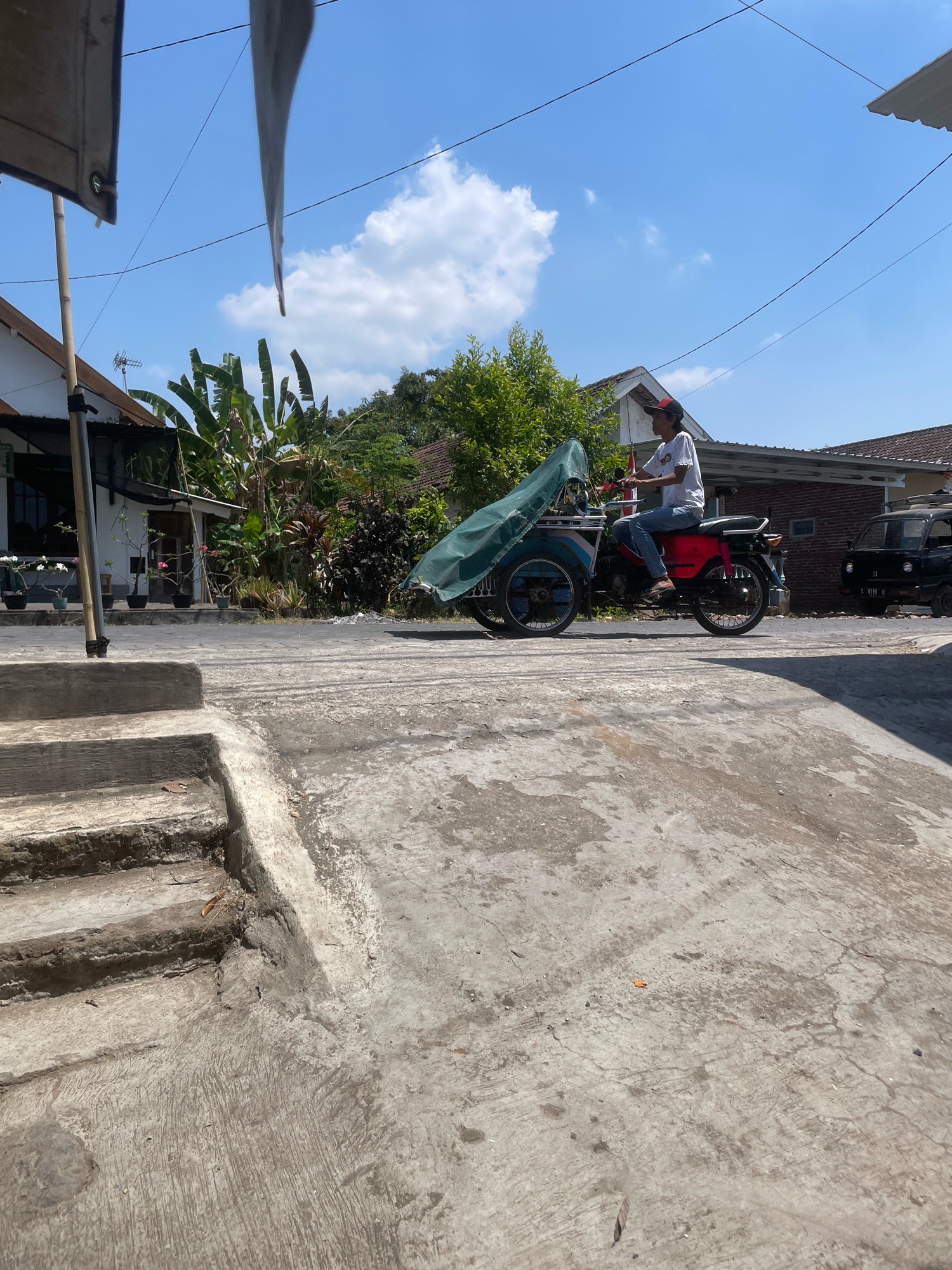 A man wearing a white t-shirt, blue jeans, and a red and black baseball cap rides a red and blue motor-tricycle with a green tarp covering the cargo area. The tricycle has two wheels at the front and one at the rear. The man is facing left and appears to be in motion. The scene is set in a street in Kebon Waris, Indonesia, with houses and lush green trees visible in the background under a clear blue sky with white clouds. The foreground features a concrete walkway with steps. A black vehicle is parked on the right side of the street. The overall impression is a sunny day in a semi-urban environment.