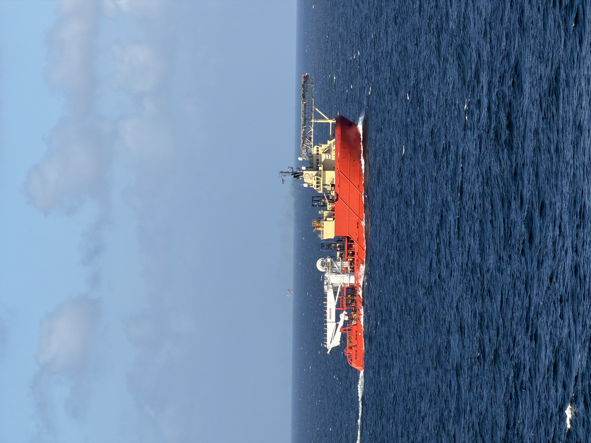 The image shows a large, red and white ship sailing on a choppy, dark blue sea. The sky is light blue with some white clouds. In the distance, there is a very faint, small structure on the horizon. The ship is moving through the water, creating a white wake. The location appears to be the open ocean, with no land visible.