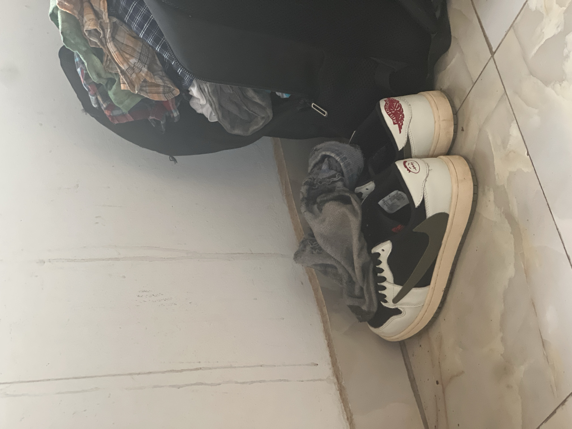 An overhead, vertically-oriented shot captures an indoor scene in what appears to be a residential setting in Abuja, Nigeria. The primary subjects are a pair of sneakers, a black backpack, and various articles of clothing, resting on a tiled floor next to a white wall.
The sneakers are prominent, positioned side-by-side with their toes facing towards the lower right of the frame. They are high-top shoes, featuring a black, white, and muted olive green color scheme, notably an inverted olive green Nike swoosh on the outer side. The shoe on the left (closer to the wall) displays a red Air Jordan "Wings" logo on its ankle collar. The shoe on the right features a red inverted smiley face, also known as the "Cactus Jack" logo, on its ankle collar, indicative of a Travis Scott collaboration. Partially visible text on the side of one shoe includes "AIR JORDAN" and "NIKE" on a tongue label. The soles of the sneakers are an off-white or cream color, showing some signs of wear or discoloration. Gray socks are crumpled inside the sneakers.
Behind the shoes, a black backpack is visible, appearing to be slightly open. Inside the backpack, various items of clothing are loosely packed, including plaid shirts (red, green, beige, dark blue) and other solid-colored shirts or fabric. White and gray socks are also visible spilling from the backpack's opening.
The floor is covered with light-colored, possibly off-white or beige, ceramic tiles, featuring a subtle, irregular veining pattern that gives them a slightly textured, natural stone appearance. Grout lines crisscross the floor. The wall adjacent to the floor is painted a plain white, showing some faint horizontal marks or stains. No specific activity is taking place; the items appear to be casually stored or momentarily placed. The time of day and weather are not discernible, but the lighting suggests an indoor setting, possibly with natural light from an unseen window or artificial lighting. No people or overt emotional cues are present.