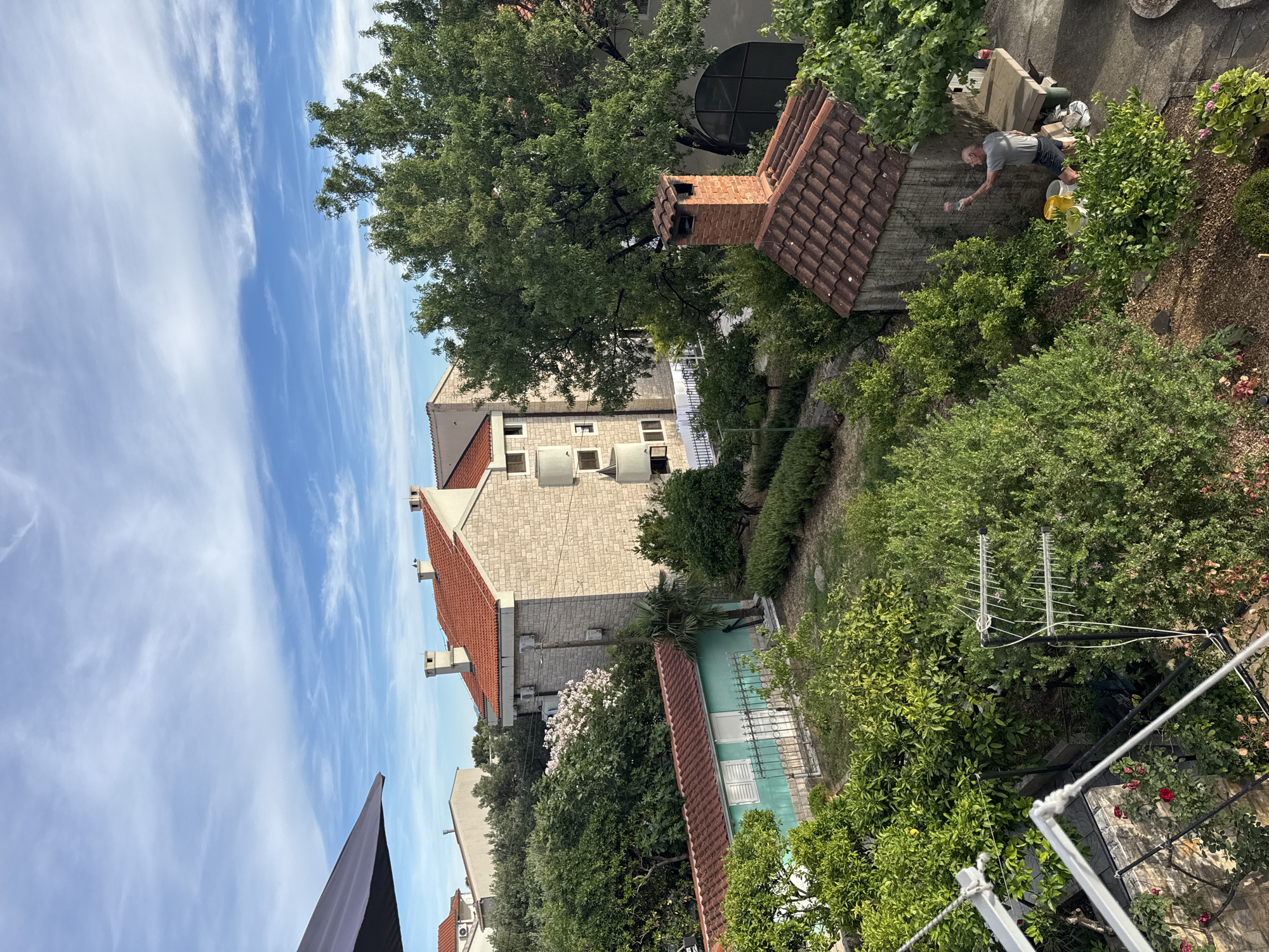 In Makarska, Croatia, on a sunny day with wispy clouds, an elderly man in a grey t-shirt and dark shorts is actively painting a small brick structure. He holds a pink paintbrush in his right hand and is reaching towards the wall. Nearby, yellow and white buckets are placed on the ground, suggesting he is in the midst of his task. The scene is set in a lush, green garden filled with trees and bushes, some bearing fruit. A terracotta-tiled roof with a brick chimney sits atop the structure he is painting. In the background, a multi-story building with light-colored stone facade and red-tiled roofs dominates the skyline. A smaller turquoise building with a red roof is also visible, bordered by a metal fence and more greenery. A large, leafy tree shades a portion of the garden. The overall impression is one of a residential area with abundant vegetation under a clear blue sky.
