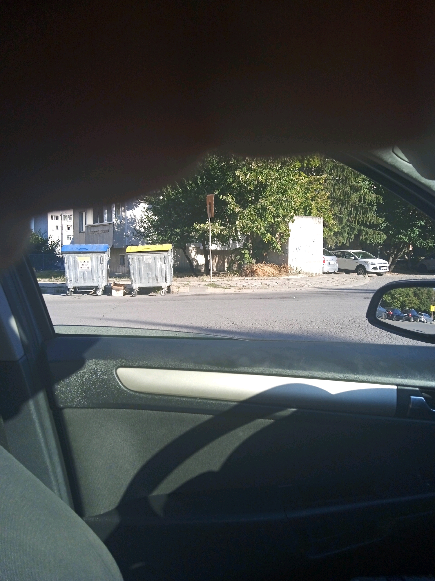 The image shows a street scene, presumably in Shumen, Bulgaria, viewed from inside a vehicle. Two large, wheeled trash receptacles, one with a blue lid and the other with a yellow lid, stand on the sidewalk near a building. A white car is parked on the street, and a white structure, possibly a wall or a utility box, is visible behind it. Trees line the background, and a side mirror of the car reflects other vehicles on the road. The interior of the car, including the door panel and a seat, is visible in the foreground.
