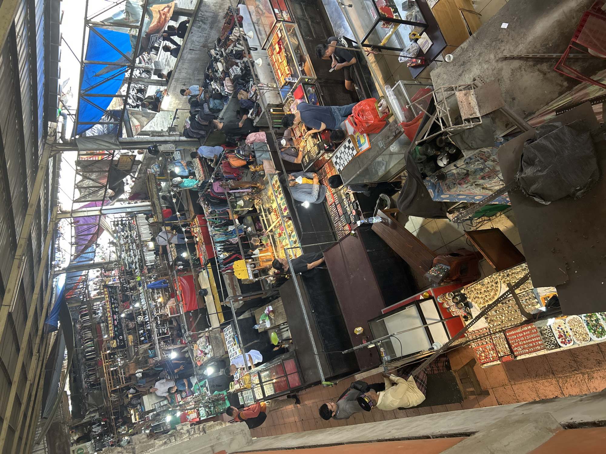 This is an overhead view of a bustling indoor market in Denpasar, Indonesia, likely a flea market or a traditional market selling various goods. The market is covered by a corrugated metal roof supported by metal beams, with tarps of blue and purple fabric stretched across some sections to provide shade. Sunlight filters through gaps, creating bright patches and shadows.
The market is densely packed with stalls and displays. In the foreground, to the left, a man in a cream-colored shirt and plaid pants sits on a wooden stool behind a display of rings and jewelry laid out on red and black trays. Next to him, a glass display case shows more rings and jewelry. Further back, on the left side of the image, a stall is filled with racks of clothing.
In the center of the image, several stalls are dedicated to jewelry and accessories. Men are seen browsing and interacting with vendors. One man in a blue shirt is looking at a display of watches. Another man in a dark shirt is showing something to a vendor. Red bags and various items are scattered on tables and on the floor.
To the right, more stalls are visible, some with clothing racks and others displaying bags and accessories. People are walking through the aisles, examining goods, and interacting. The overall environment appears chaotic but organized, with a continuous flow of activity. The stalls are constructed from wood and metal, with many displaying their wares on shelves and tables. The floor is tiled or concrete. The scene conveys a sense of commercial activity and community gathering.