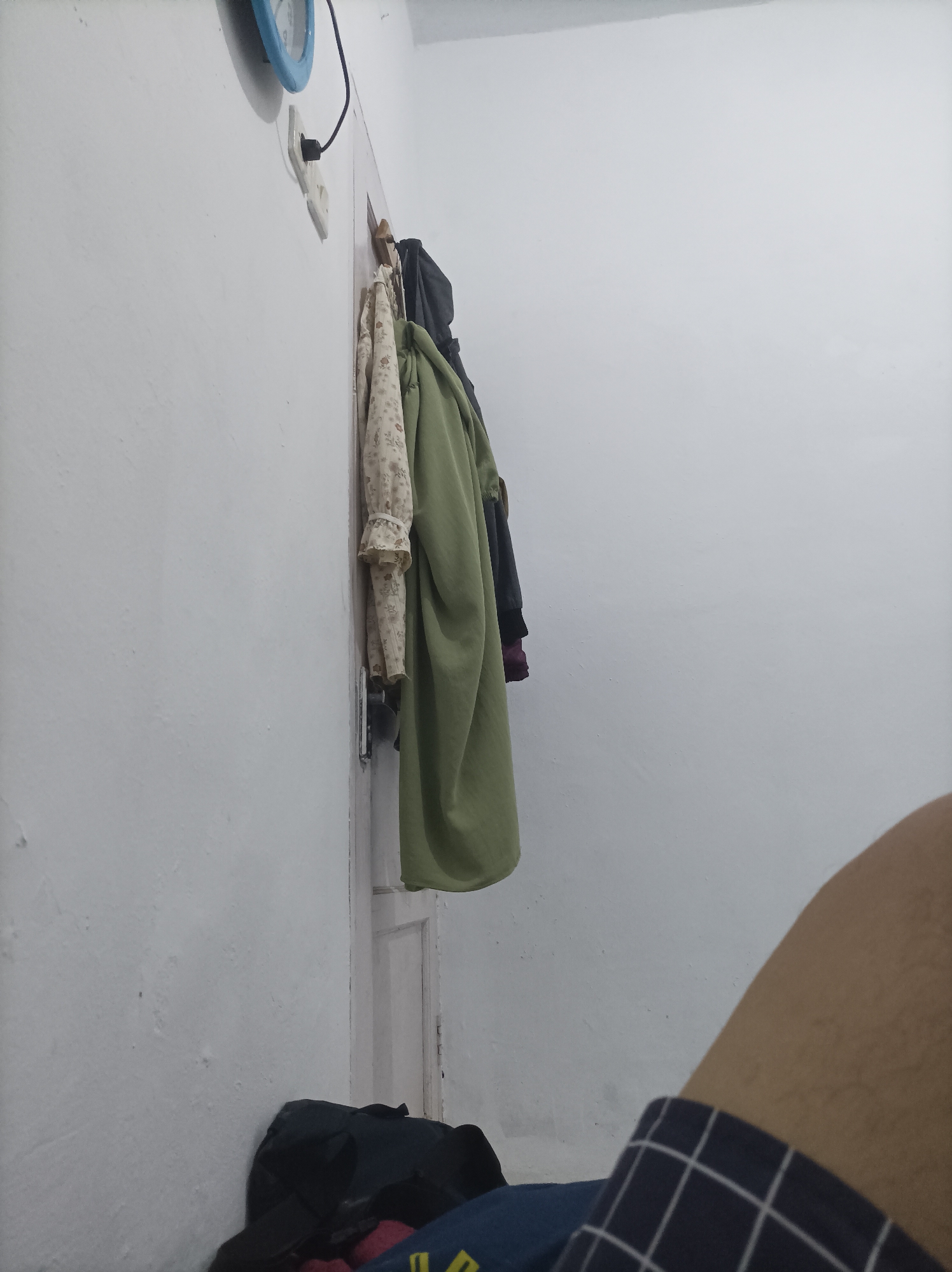 A close-up, low-angle, indoor shot shows a white wall with some texture and a light-colored door frame on the right. A blue-faced clock with black hands is mounted on the wall near the top left corner, connected to a white electrical outlet by a black cord. Several pieces of clothing hang on hooks on the door frame: a light-colored floral patterned garment, a green garment, and a darker garment. At the bottom of the frame, on the floor, is a black bag and a glimpse of a person's leg covered by plaid pajama bottoms. The scene appears to be within a residential setting in Pamengkang, Indonesia.