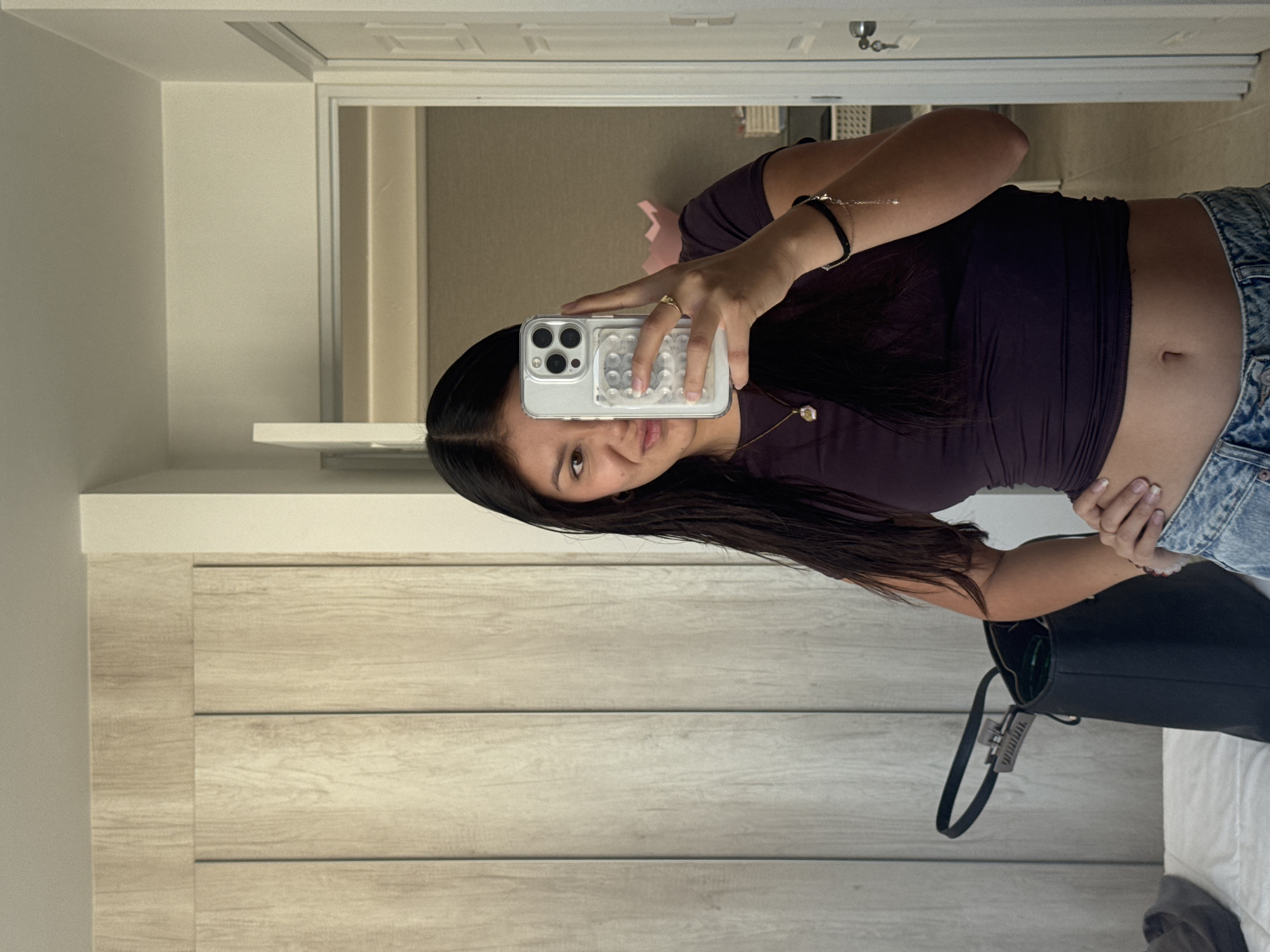 A woman is taking a selfie in a room. She is holding a smartphone with a clear case in her right hand. The woman is wearing a dark purple top and light wash jeans. She has dark hair. A black purse rests on a surface beside her. The background includes a door, light-colored wood-grain paneling, and a glimpse into another room. The specific location is not discernible from the image.