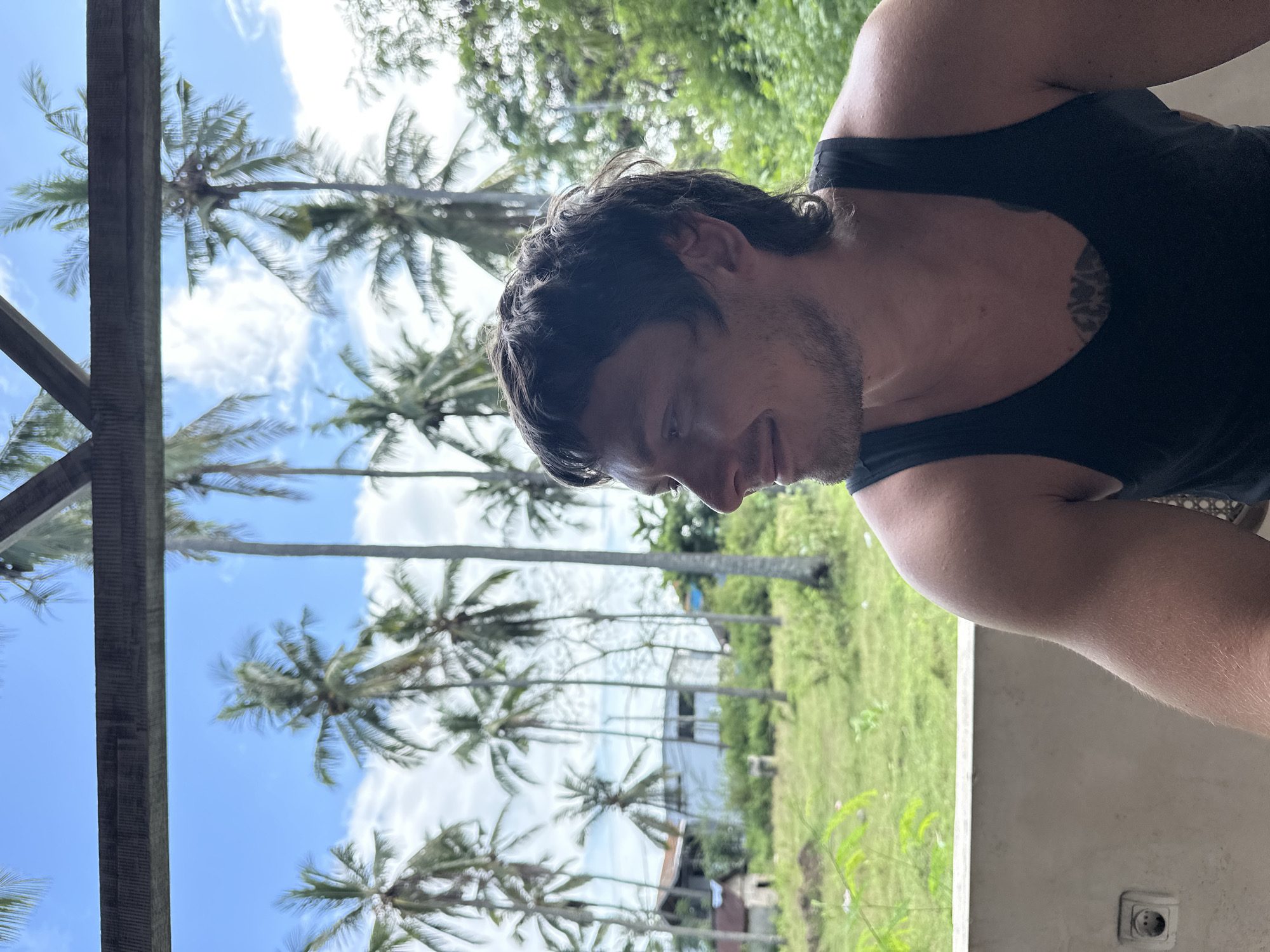 A man wearing a black tank top is situated indoors with a view of palm trees and a green landscape. The location appears to be Nusa Penida, Indonesia. The image includes elements of architecture, greenery, and a clear sky with light clouds. An electrical outlet is also visible.