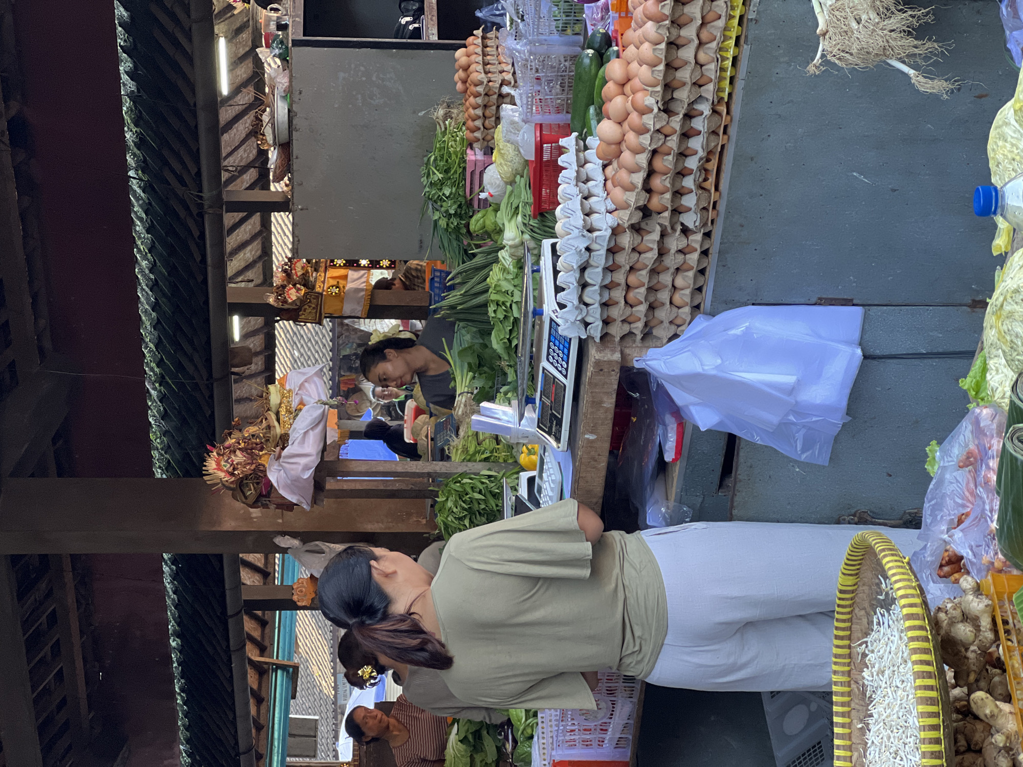 This image captures a vibrant scene at a market in the Ubud District, Indonesia. The focus is on a woman in a khaki shirt and white pants, standing near a vendor's stall laden with fresh produce and eggs.
Behind her, another woman with dark hair is visible, seemingly engaging with the vendor. A scale rests on the counter, suggesting transactions are taking place. The stall is overflowing with a variety of items, including stacks of eggs in cartons, fresh vegetables like greens and long beans, and what appears to be onions with their roots. Other items like sprouts and ginger can be seen, contained in baskets and plastic packaging.
The market's architecture is characterized by a rustic, traditional structure with wooden beams and roofing. In the background, there are details that suggest offerings or decorations, indicative of Balinese Hindu practices. The overall impression is of a bustling marketplace in a traditional Indonesian setting.
