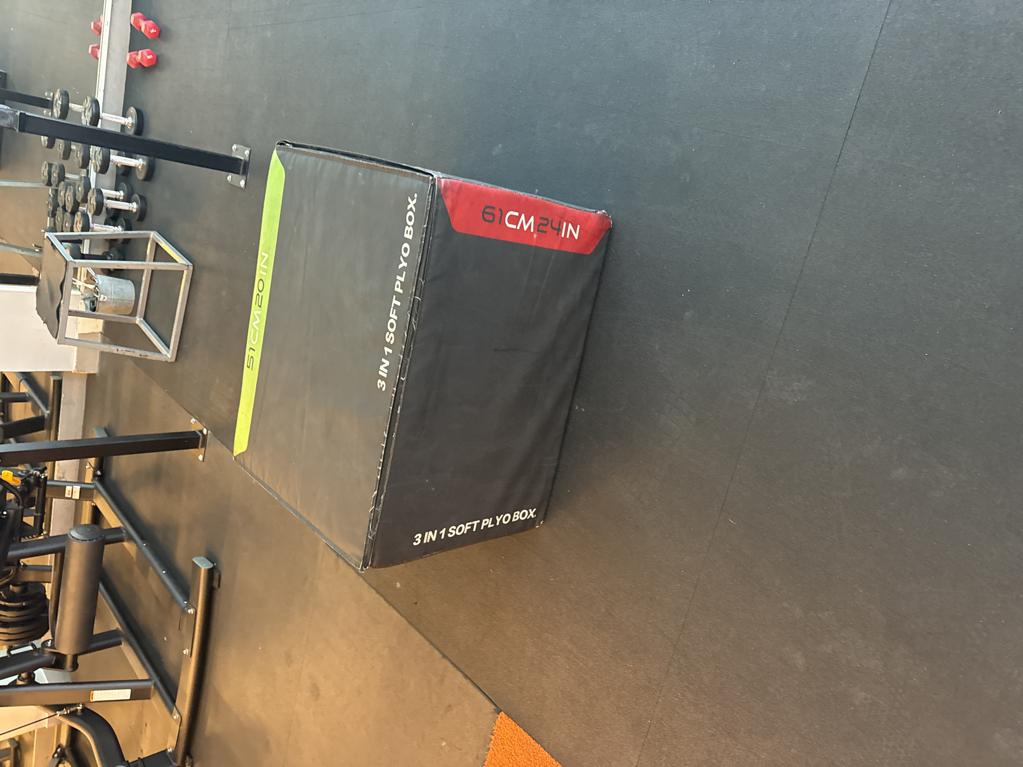 The image shows a gym setting with various exercise equipment, including dumbbells and a plyometric box. The plyo box, which appears to be made of soft material, is black with text labels in white and red. One label reads "51CM 20 IN", and another says "3 IN 1 SOFT PLYO BOX". A red label on the corner of the box shows "61CM 24IN". In the background, metal racks hold sets of dumbbells, and red-colored weights are visible on the floor. The gym floor is dark, likely a rubberized matting common in fitness facilities. The lighting suggests it's daytime, and the environment appears to be indoors. No people are visible in the frame, focusing the attention solely on the equipment. The scene is static, capturing a typical arrangement of fitness gear ready for use. The image is oriented vertically.