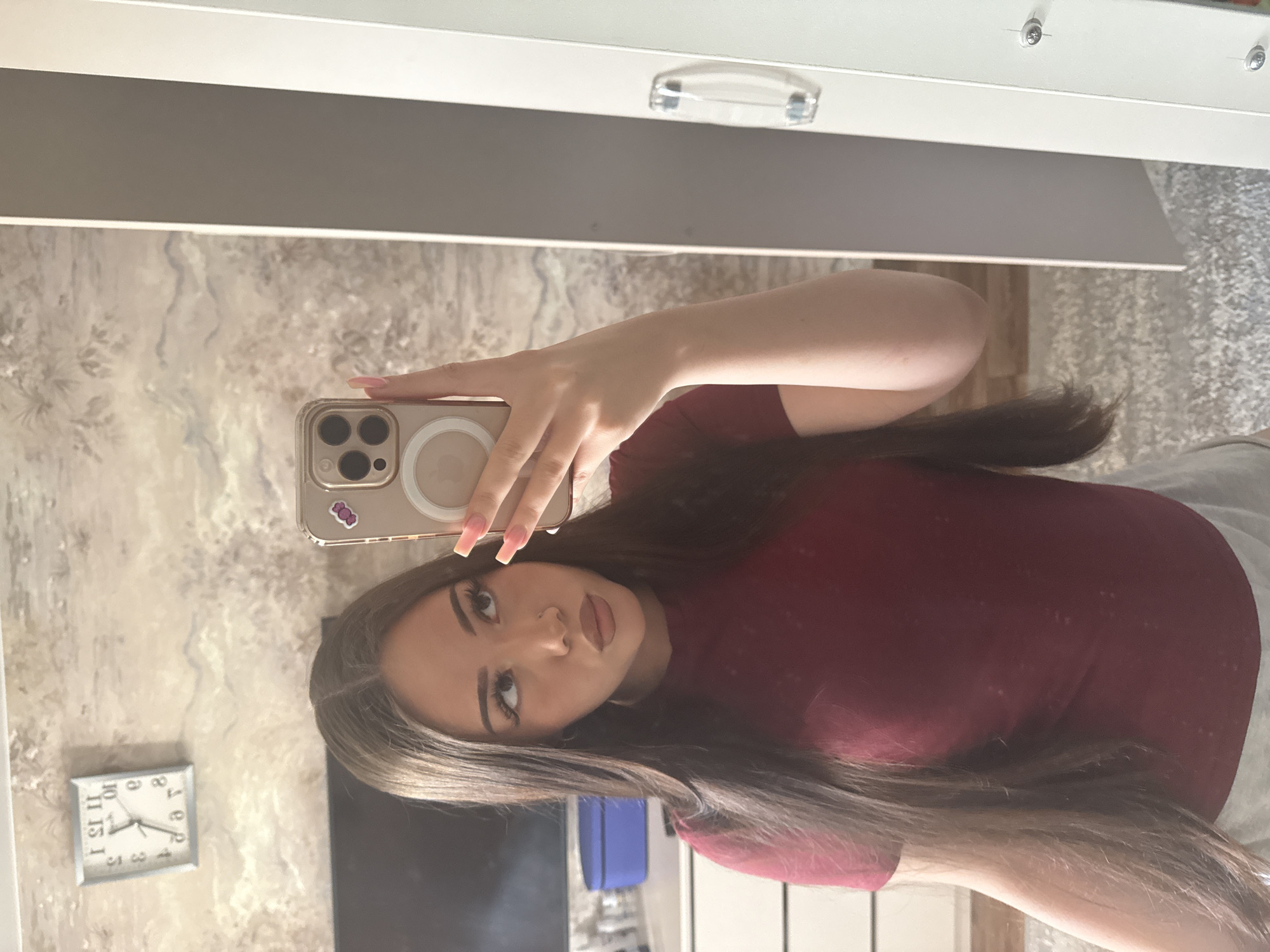 An indoor mirror selfie captures a young woman holding a smartphone. The image is oriented vertically (rotated 90 degrees counter-clockwise from its original capture). The woman, likely a young adult, has long, straight dark brown hair. She is wearing a maroon or burgundy short-sleeved top. Her makeup is visible, featuring defined eyebrows, eyeliner, and contoured lips. She has long, light pink/nude colored nails. Her expression is neutral to slightly alluring.
She is holding a smartphone, specifically an iPhone with a prominent three-lens camera module and a white MagSafe ring visible on its clear case. A small sticker is affixed to the back of the phone case, though its design is unclear.
The setting appears to be a room, possibly a bedroom or living area in Naberezhnye Chelny, Russia. The background features light-colored, subtly textured wallpaper or wall covering. Above the woman's head (when viewed upright) is a white piece of furniture, likely a shelf or cabinet, with a clear handle. To her left (when upright), a square wall clock is reflected on the wall. Its numbers and hands are mirrored. The clock's hands, as seen in the reflection, indicate approximately 11:05 (the hour hand pointing to the mirrored '11' and the minute hand pointing to the mirrored '1'). This corresponds to an actual time of roughly 1:55. A fluffy, light-colored carpet is visible to the right of the woman's arm. The lighting suggests daytime.