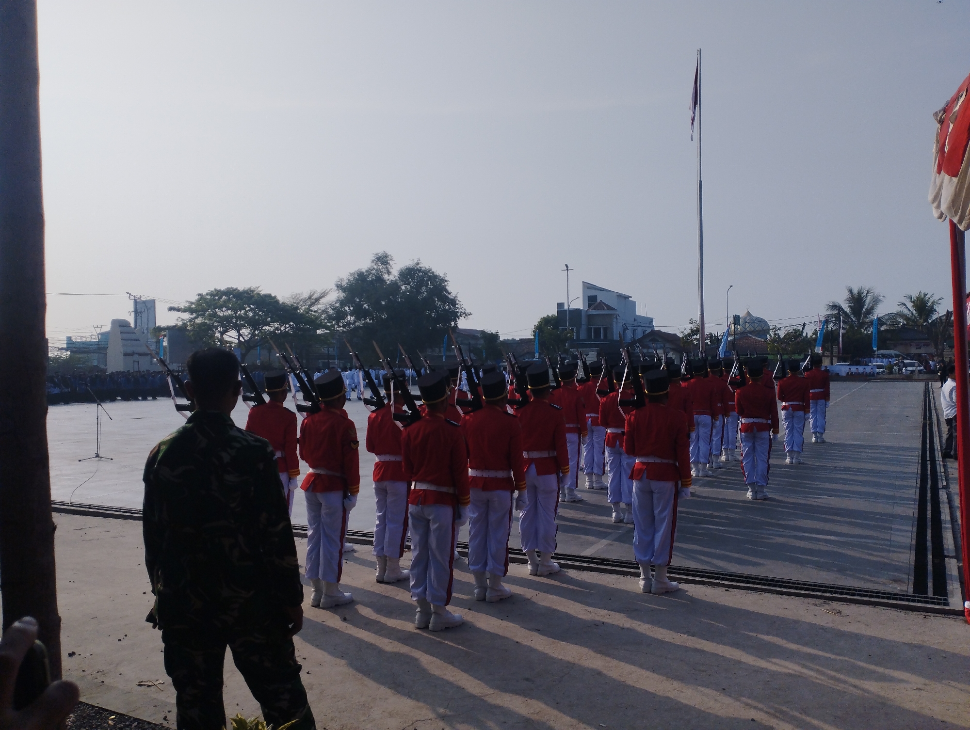 The image depicts a clear, overcast day in Kramatwatu, Indonesia, possibly in a public square or training ground. In the foreground, a person in camouflage fatigues stands with their back to the camera, facing a formation of individuals in bright red ceremonial uniforms with white pants and black hats. These individuals are standing in rows, holding rifles horizontally. The formation is marching or preparing to march on a paved surface. In the background, buildings, trees, and a flagpole with a flag are visible, suggesting an urban or semi-urban environment. The overall scene appears to be a military or ceremonial event.