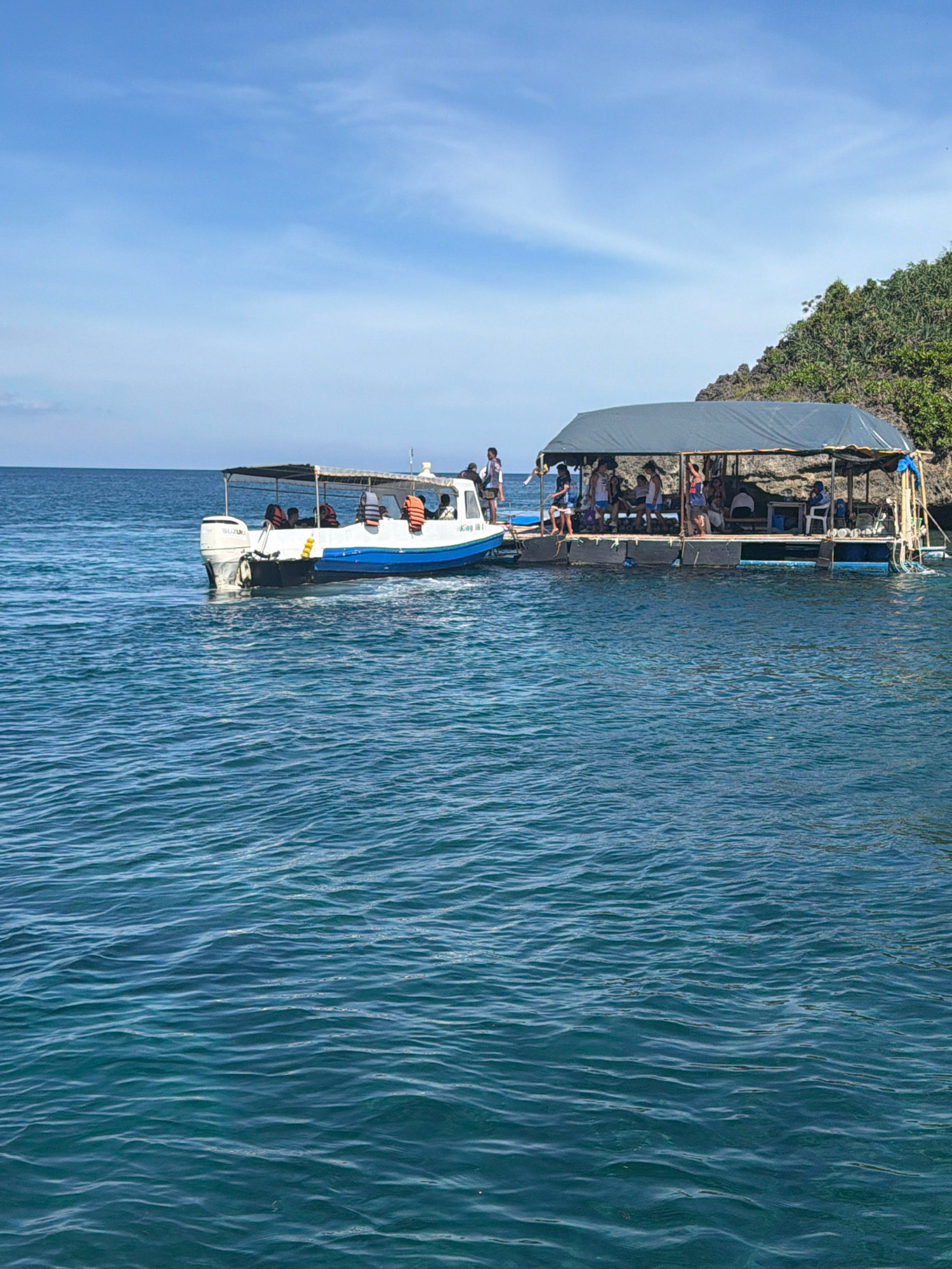 A small motorboat, with a blue and white hull and a white canopy, is moored alongside a floating platform covered by a dark grey canopy. The platform is crowded with approximately 15 people, both men and women, some standing and some seated on benches. A few individuals are visible on the boat as well, some wearing life vests. The scene is set in a body of clear, blue water, with a rocky, tree-covered island in the background. The sky above is a bright, clear blue with a few wispy clouds. The location is identified as Bakhaw, Nabas, Philippines.