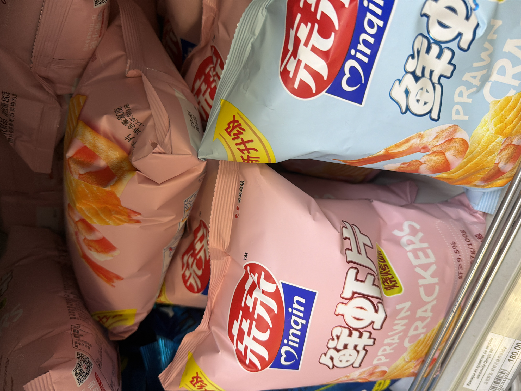 The image shows a close-up view of several bags of "Qinqin" brand prawn crackers, likely on a store shelf. The bags are pink and light blue, featuring images of prawns. Text on the packaging is a mix of English and Chinese characters. The packaging also indicates that the crackers are shrimp flavored. The lower-right corner contains Russian text and a price, suggesting a location in Russia, most likely Moscow.