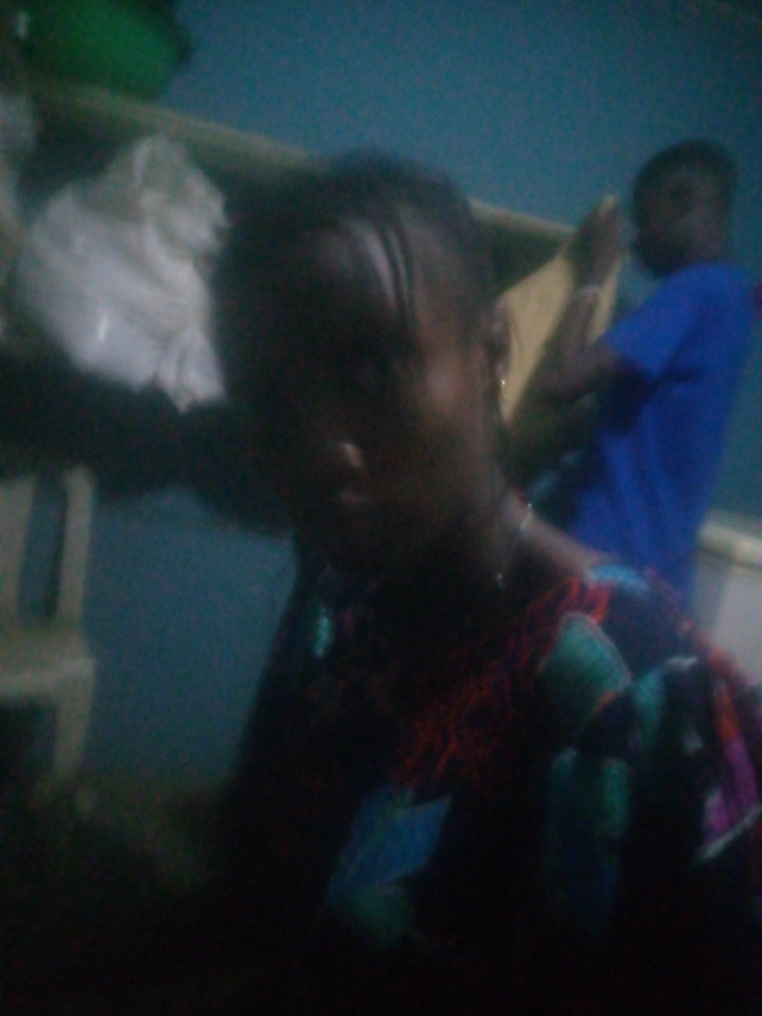 Here is a detailed description of the image:
The image shows two people in an indoor setting. The person in the foreground is partially visible, wearing a colorful patterned top. The other person is further back, wearing a blue shirt. The setting appears to be a room with a blue wall. Some objects such as a white bag on a chair and other indistinct elements are visible in the background. The location is Unknown, Unknown.