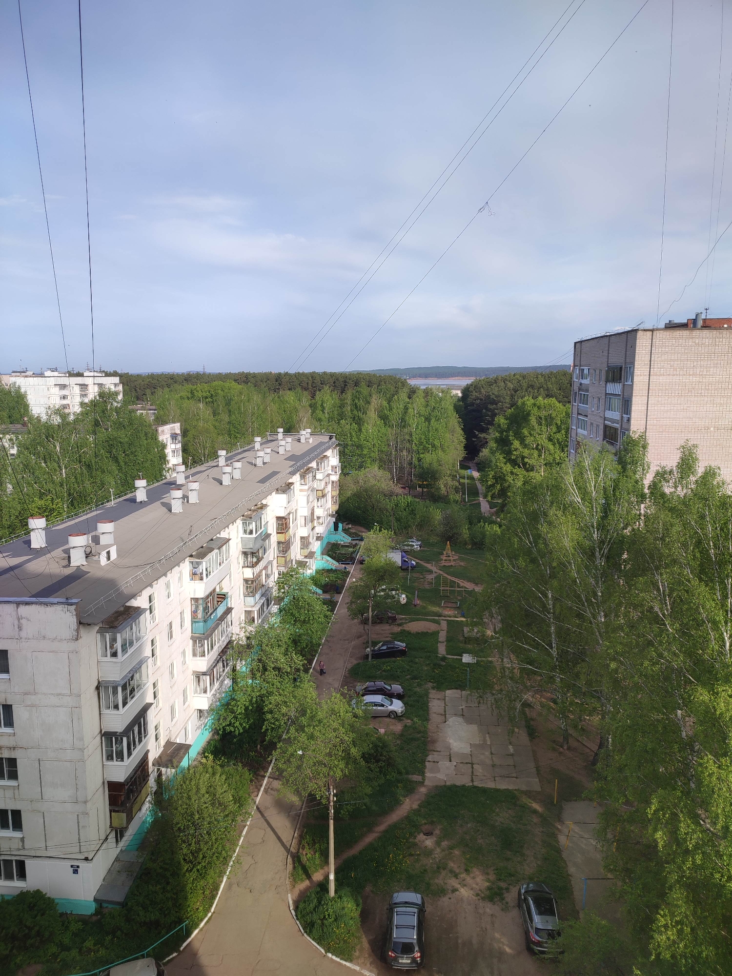 The image depicts a residential area within the city of Unknown. A multi-story apartment building with a gray roof is visible on the left side of the frame. Additional apartment buildings, surrounded by dense trees, are present in the background. A courtyard area, featuring patches of grass, trees, and a walkway, is situated between the buildings. Several cars are parked in designated parking spots within the courtyard. Power lines are visible stretching across the top portion of the image. The sky is partly cloudy.
