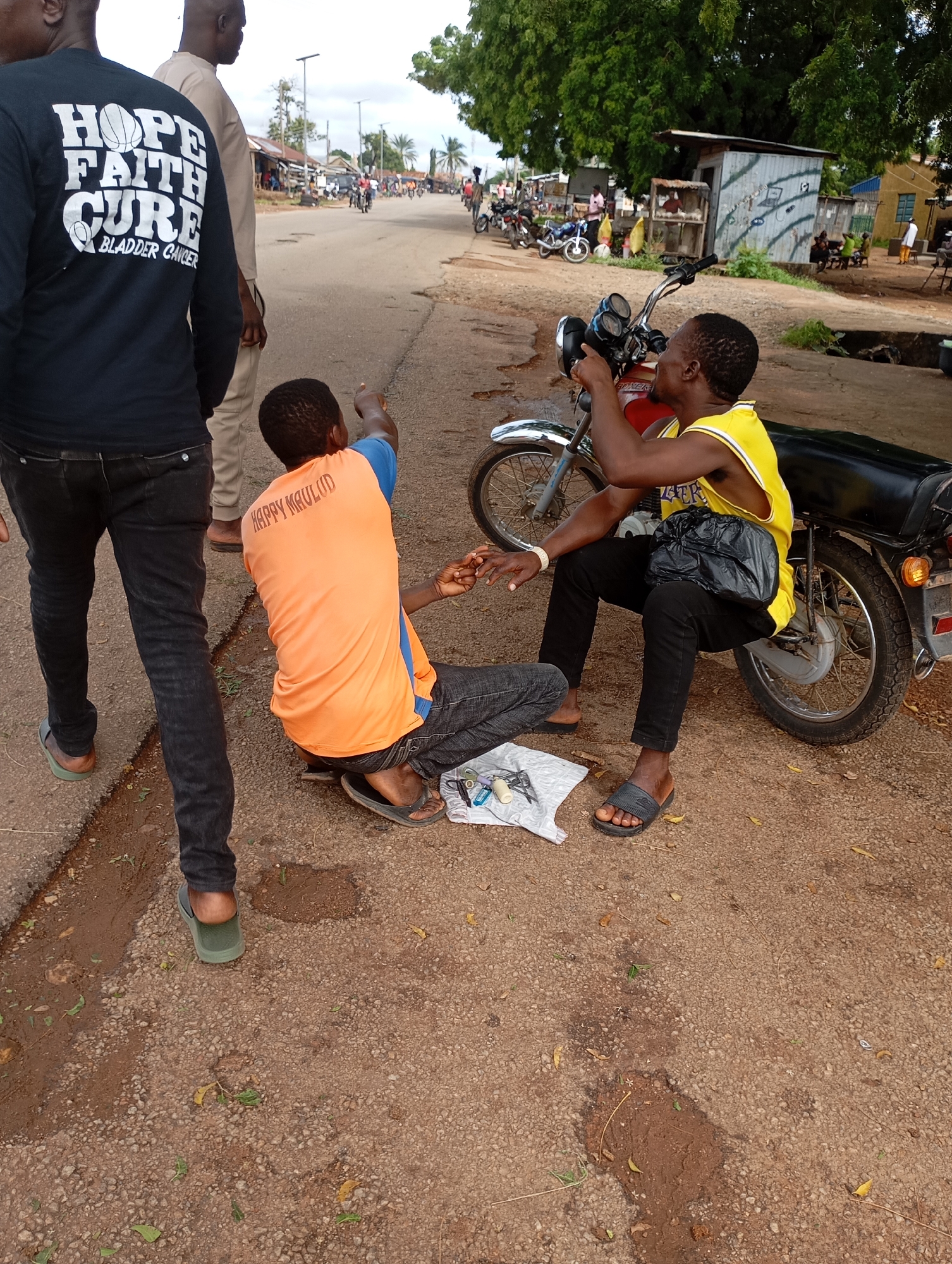 In Shendam, Nigeria, two young men are gathered on a dusty roadside next to a red and black motorcycle. One man, wearing a black long-sleeved shirt with white lettering that reads "HOPE FAITH CURE BLADDER CANCER," stands and points towards something off-camera. The other man, seated on the ground, is wearing an orange t-shirt with blue trim and has the words "HAPPY MAULUD" printed on the back in orange. He is looking down at a small white bag containing various items, possibly tools or parts, and appears to be working on something near the motorcycle's wheel. A third man, partially visible in the background wearing a beige traditional outfit, stands behind the man in the black shirt. The environment is characterized by a paved road, a dirt shoulder, and sparse vegetation, with buildings and trees lining the street in the distance.