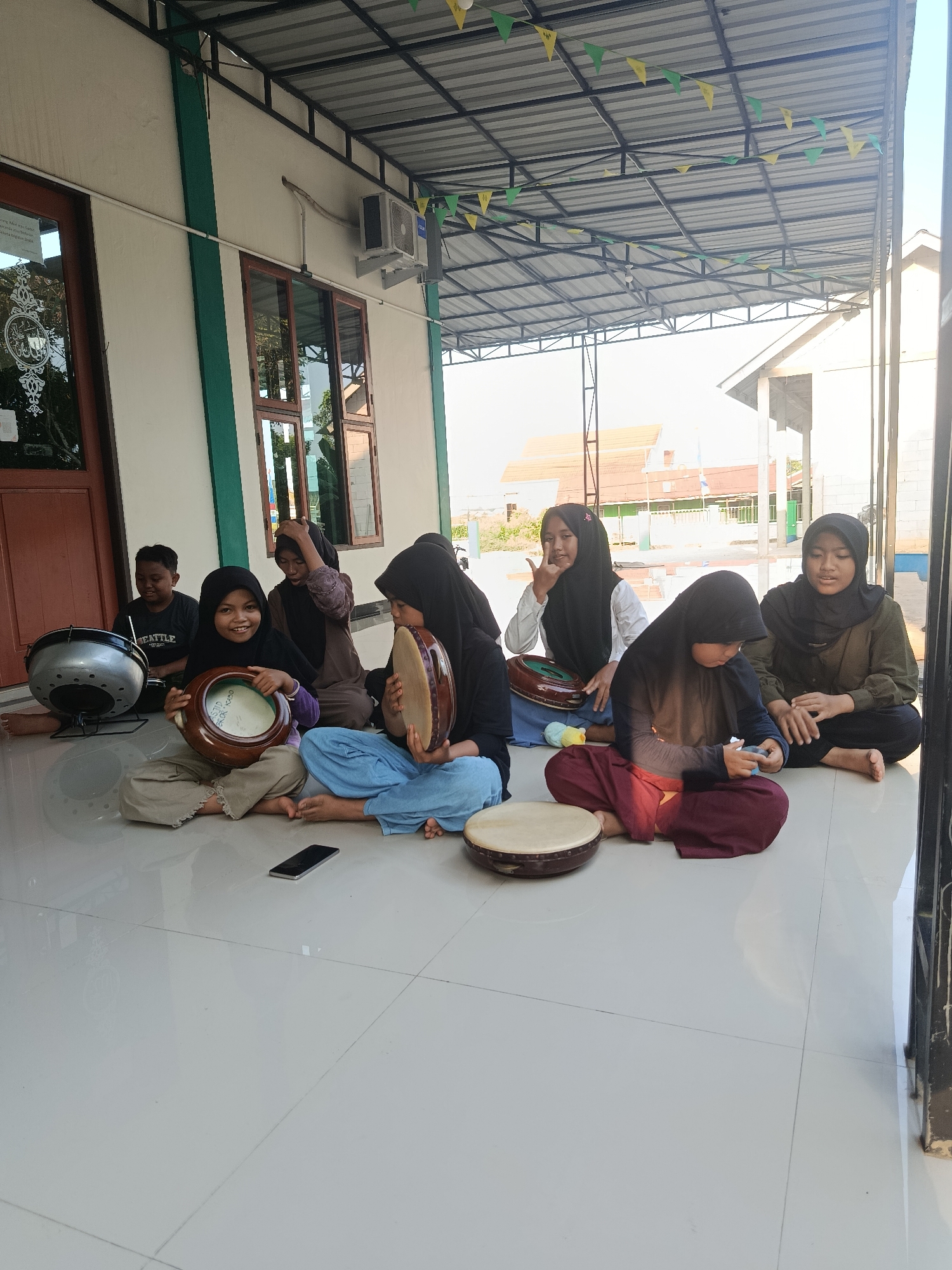 In Swarga Bara, Indonesia, a group of children, mostly girls wearing hijabs, are seated on a tiled floor outside a building with a metal roof. They are holding and playing hand drums. One girl in the foreground is smiling at the camera, holding a drum. Another girl next to her is looking down at a smartphone. To the left, a boy is looking at a large silver drum. Decorative flags are strung across the underside of the roof. The building appears to be a mosque or religious school, with large windows and a green and white painted exterior.