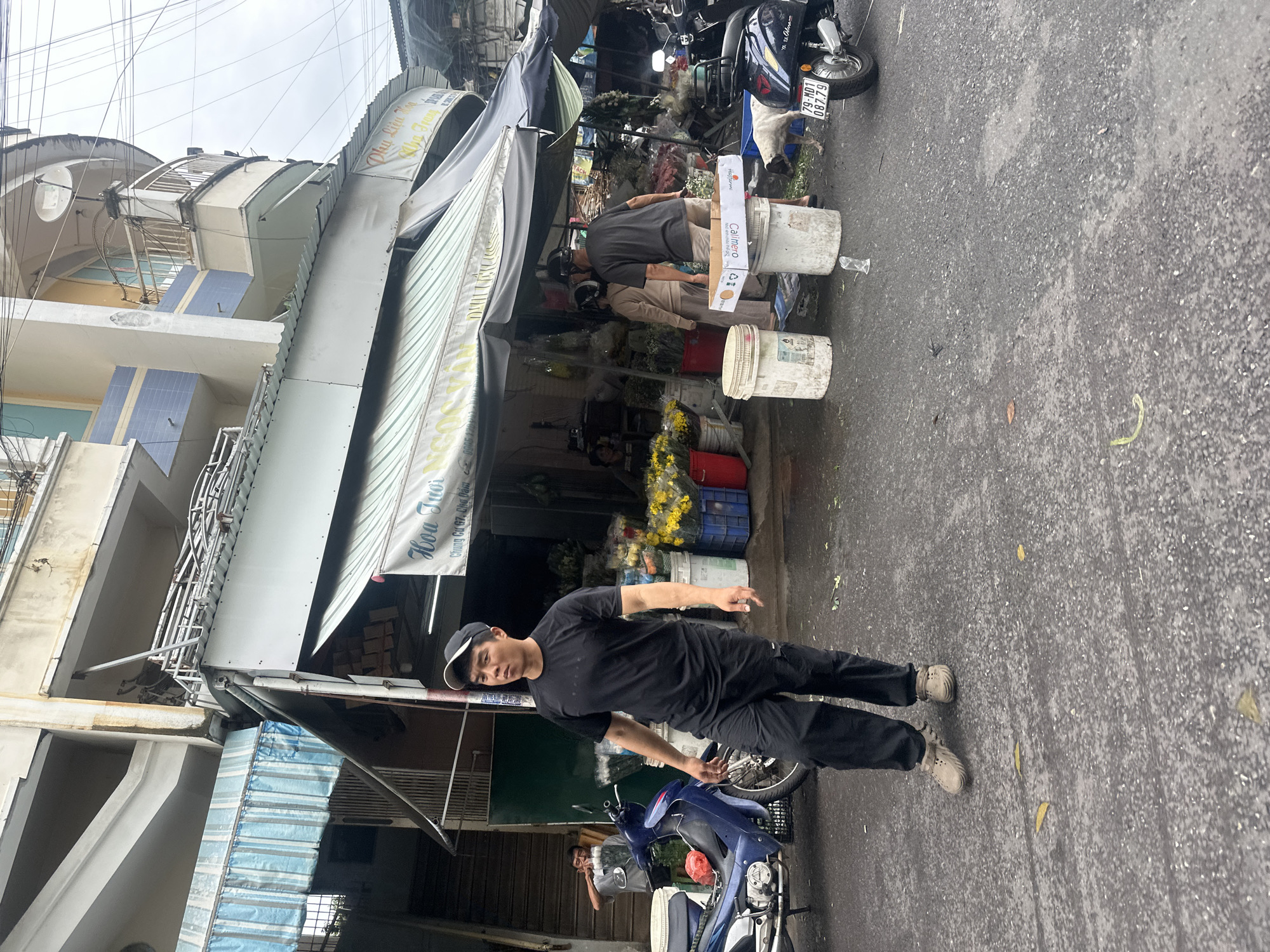 The image captures a street scene in Nha Trang Ward, Vietnam. A man in a black t-shirt, black pants, a black cap, and light-colored shoes is standing on the right side of the frame. Beside him is a parked blue scooter.
Behind him, there's a flower shop under an awning with a sign that reads "Hoa Tươi Chung Cư G7, Chợ Đầm." Two people appear to be working at the shop, arranging flowers in buckets. There are multiple white buckets filled with flowers and various colored buckets. The shop extends under the overhang of a building, with several stories visible above.
Parked next to the flower stall is another scooter, partially visible. The street surface is asphalt and appears to be wet. Power lines crisscross above the buildings. Boxes with the name "Calimero" are on the buckets. The license plate on the scooter is "79-MD1 087.79".