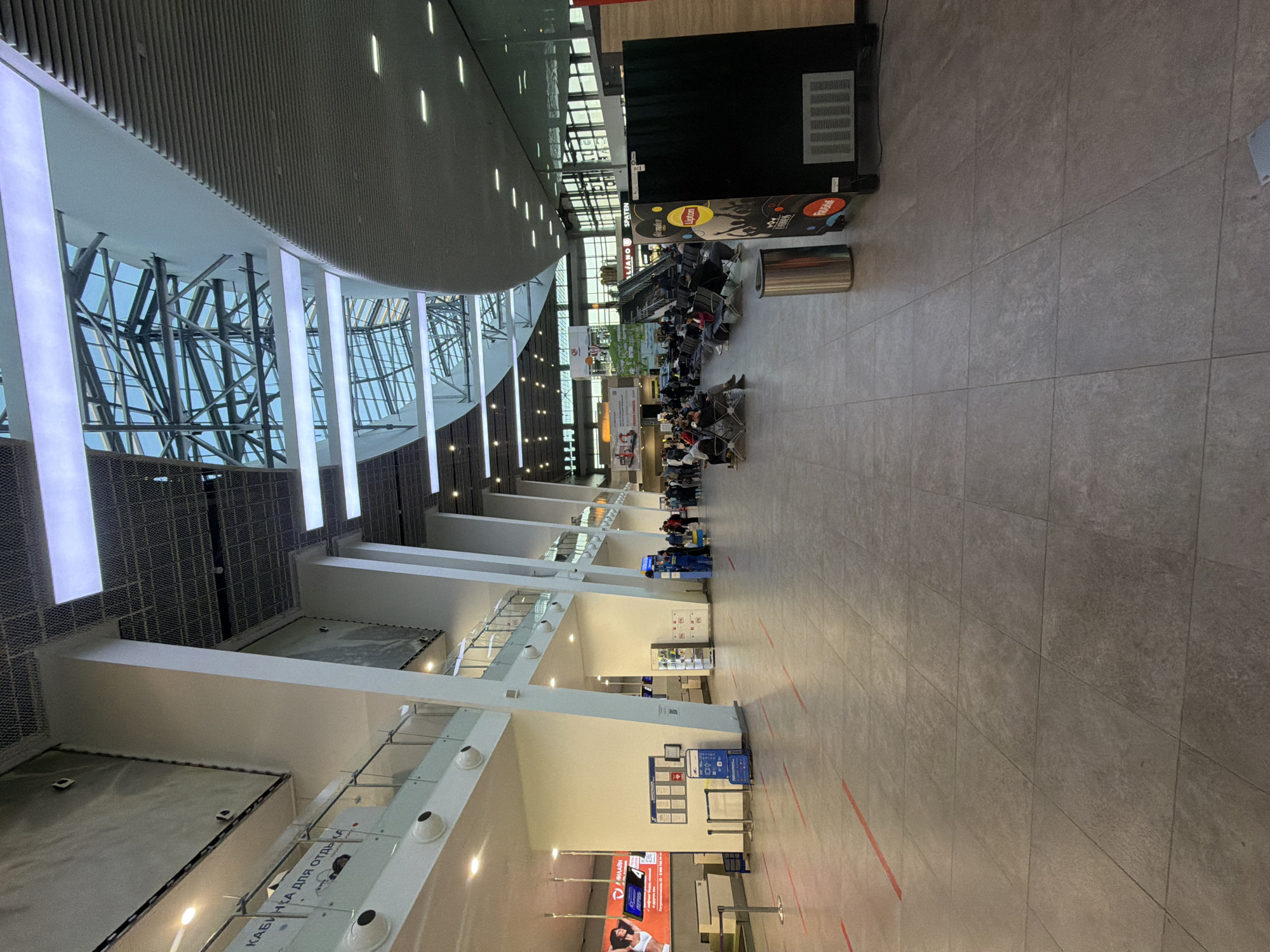 The image depicts an interior space in Bolshoye Savino, Russia, presumably at an airport or transportation hub, given the presence of seating areas with people waiting. The space is characterized by modern architecture, featuring a curved ceiling with exposed structural elements and large windows. There are rows of seats occupied by individuals, suggesting a waiting area or a queue for a service. There are also kiosks or vending machines. The floor is tiled.