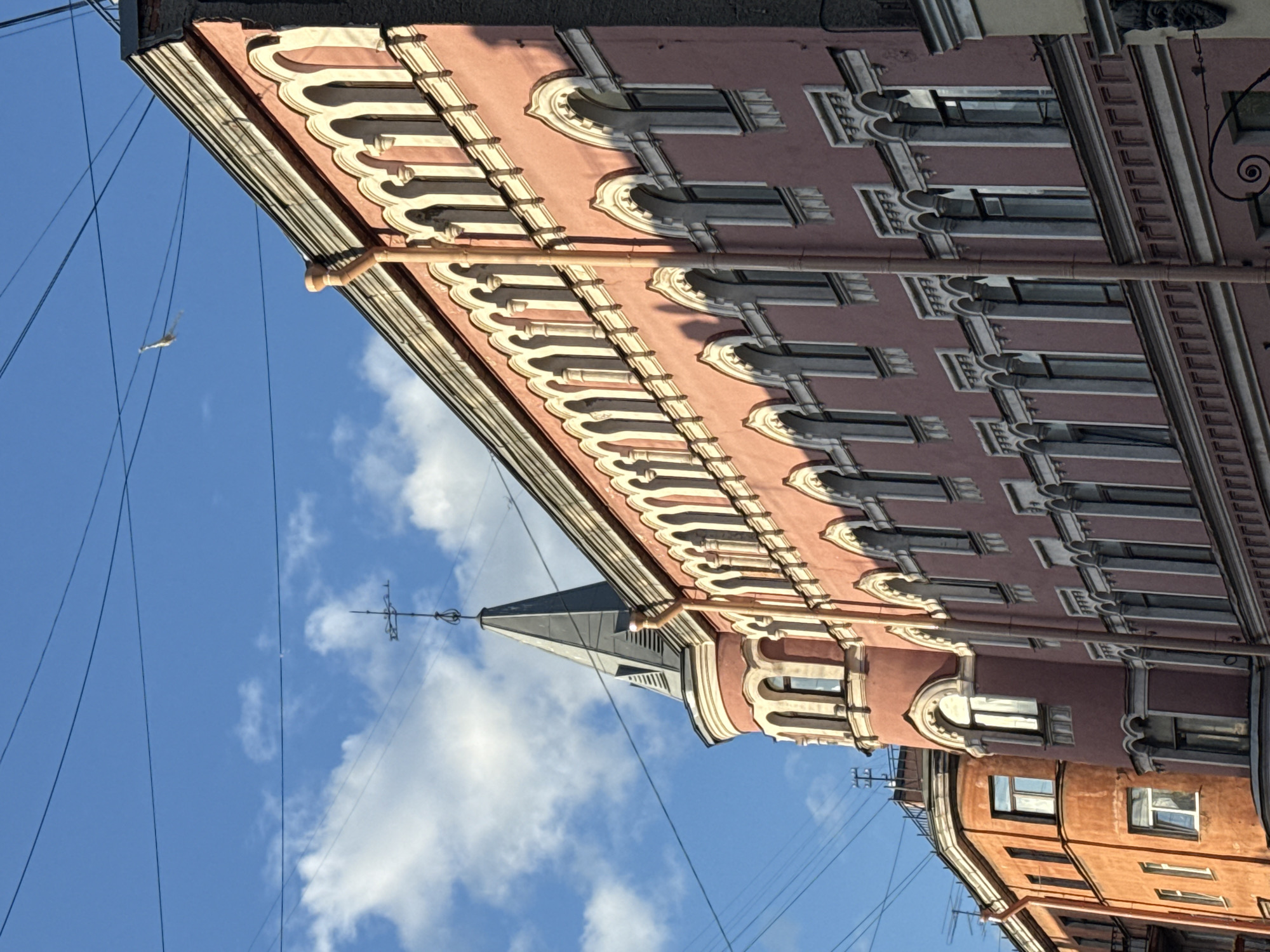 This is a photograph of an old building in Saint Petersburg, Russia, taken from a low angle. The building is painted a dusky pink and features ornate white architectural details around its arched windows and along the roofline. A conical tower with a weather vane tops the building on the left side. The sky is a vibrant blue with scattered white clouds. Overhead, power lines are visible, and a small, distant object, possibly a bird or a kite, is in the sky. To the left of the pink building, a portion of an adjacent orange building is visible, with its own distinct window patterns. The sunlight casts shadows on the pink facade, highlighting the architectural embellishments.