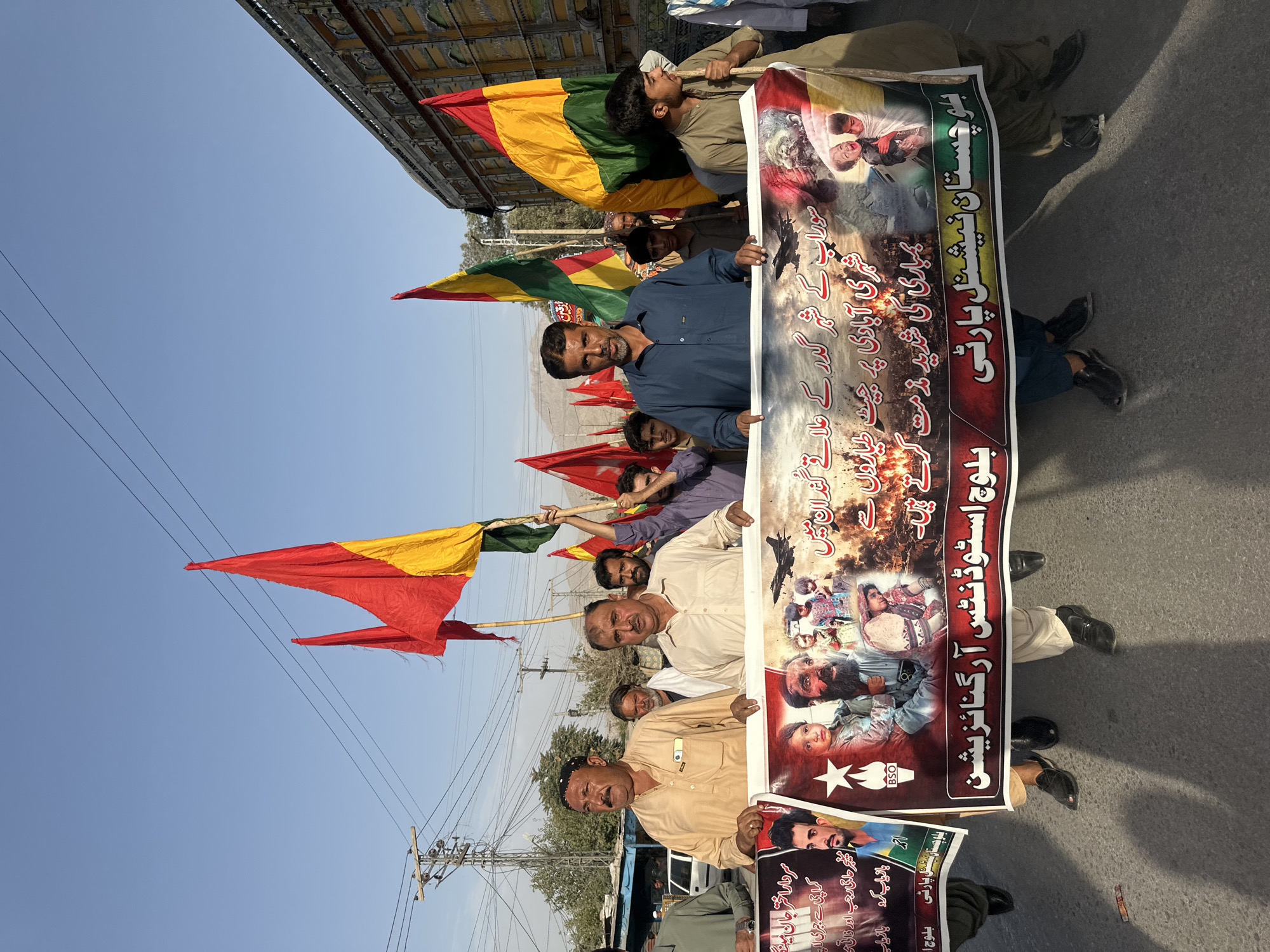 A group of men are marching outdoors in a street, holding a large banner and several flags. The banner, which appears to be made of fabric, is predominantly white with text and images in various colors. On the left side of the banner, there are portraits of men, one in a traditional Pakistani cap and light-colored shirt, and another with a beard wearing a similar shirt. A star symbol is also visible. The central part of the banner features images of children and adults, with a dark background that suggests conflict or destruction. The right side of the banner contains more text and images. The flags being held are large and rectangular, with some appearing to be in red, yellow, and green colors, possibly representing political or cultural affiliations. The men are dressed in a mix of traditional and casual attire. Some are wearing shalwar kameez, while others have on shirts and pants. The environment appears to be urban, with a street surface, overhead power lines, and some buildings or structures visible in the background. The sky is clear and blue, indicating daytime. The overall scene suggests a protest or a public demonstration. The text on the banner is in Urdu and Balochi script. The location is specified as Quetta City Tehsil, Pakistan.