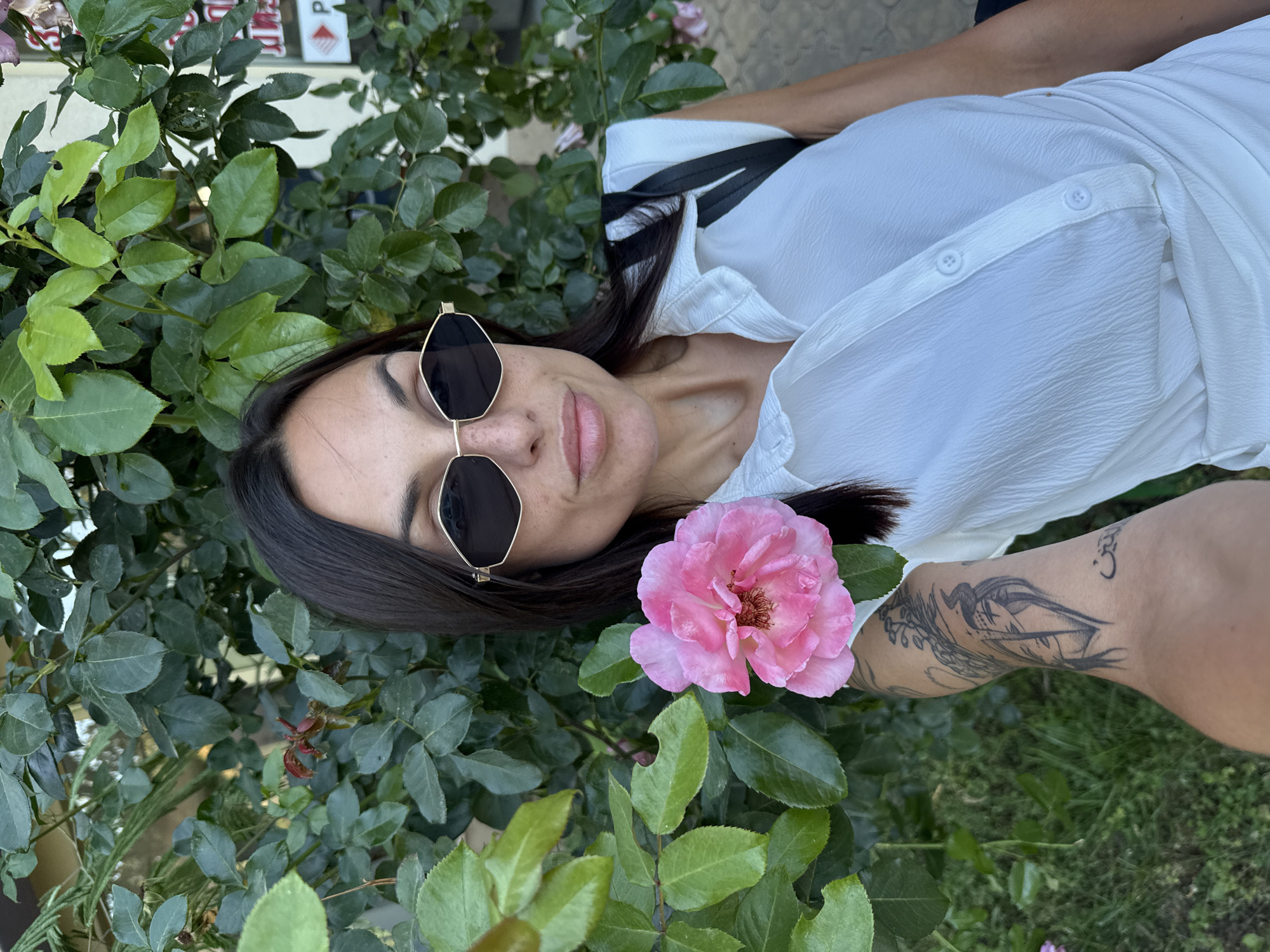 A woman with long dark hair, wearing sunglasses and a white collared shirt, is taking a selfie outdoors. She is positioned in front of a rose bush with a pink rose bloom in the foreground. A tattoo is visible on her left arm. In the background, there is a building with signage that includes Cyrillic script, suggesting a location in Pleven, Bulgaria. The overall environment appears to be an urban setting with greenery.