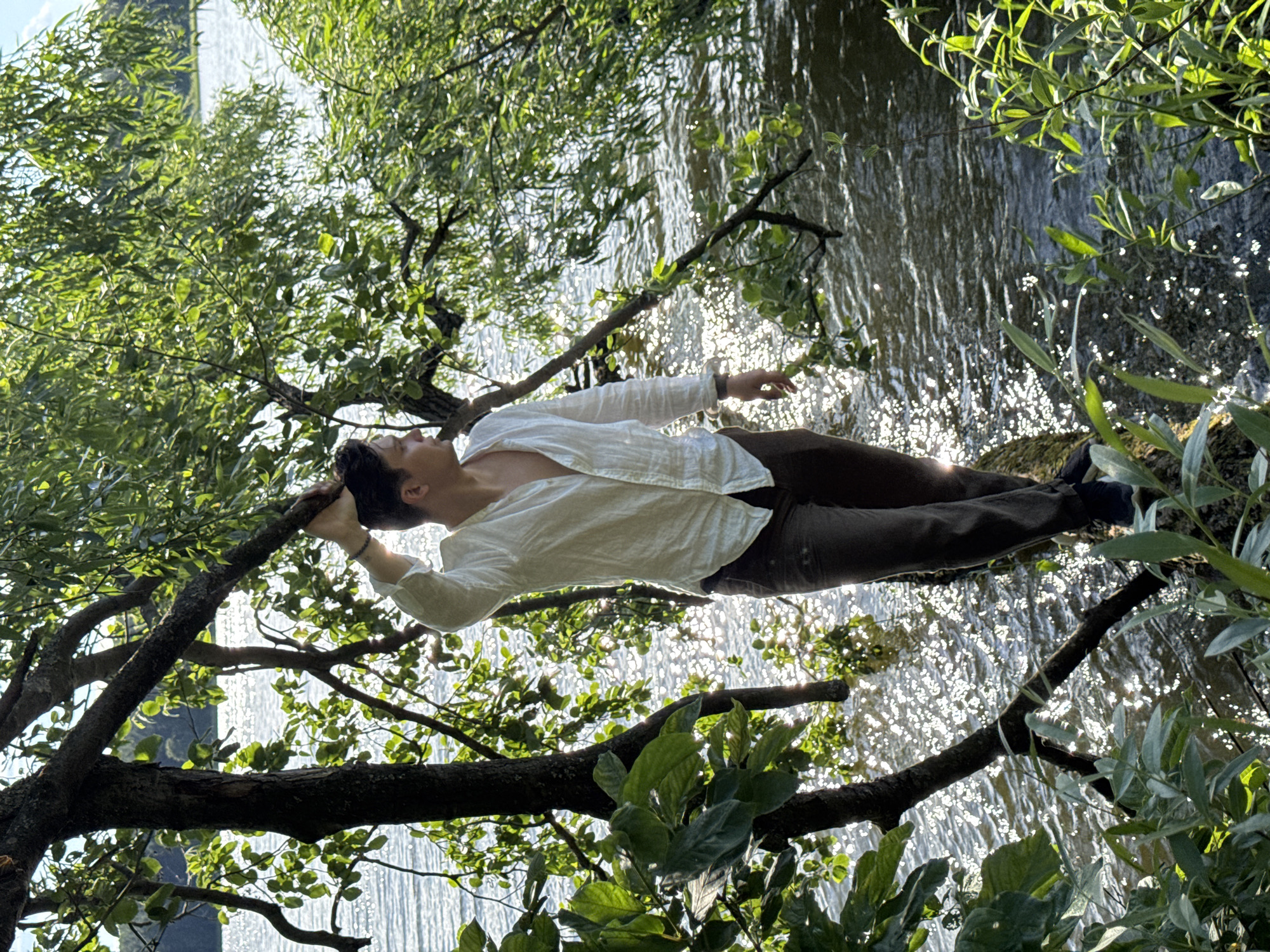 The media file depicts a young person lying on a tree branch that extends over a body of water. The person is wearing a white, loosely buttoned shirt and dark pants. Their head is resting on their hands, and their eyes are closed, suggesting a state of relaxation or contemplation. The water below is dappled with sunlight, creating a shimmering effect. Lush green foliage surrounds the scene, with the leaves of the tree creating a canopy and framing the view of the water. The lighting suggests it is daytime, likely late afternoon or early evening, given the warm tones and the angle of the sun. The scene appears to be set in a natural environment, possibly a park or a lakeside area. The overall mood is tranquil and serene. There is no visible text in the image. The location context provided is Votkinsk, Russia.