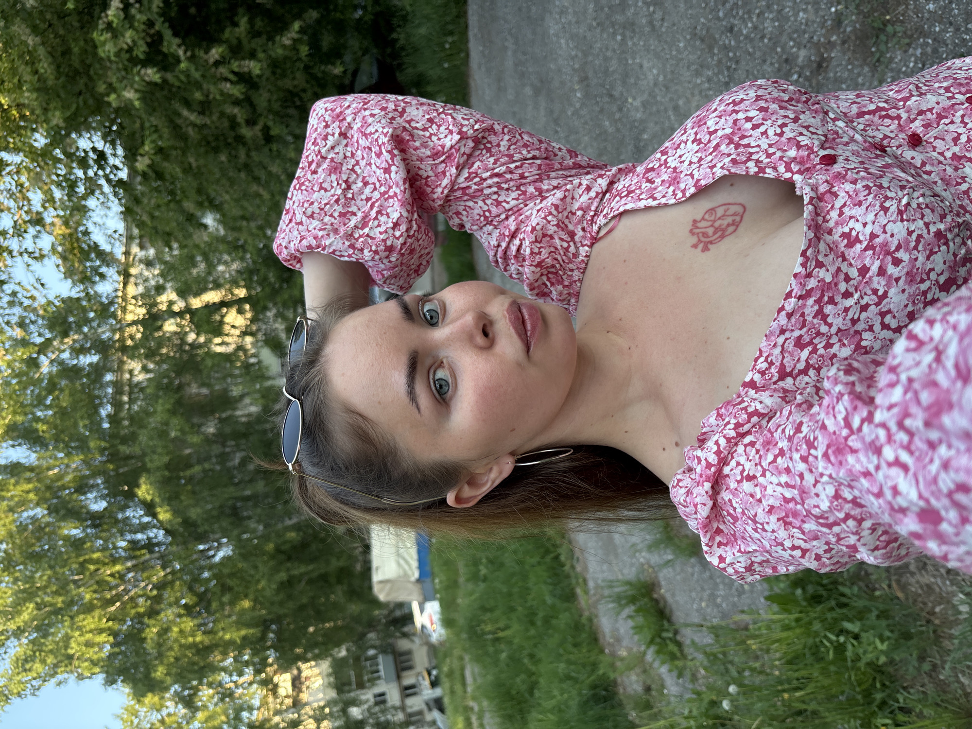 A young woman is lying down in what appears to be a park in Izhevsk, Russia. She is wearing a pink floral dress. She has brown hair, blue eyes, and is wearing hoop earrings. A pair of sunglasses is perched on her head. The woman has a tattoo on her chest. The background consists of trees, grass, and a concrete path. Buildings are visible in the distance. The image appears to be taken from a slightly elevated angle.