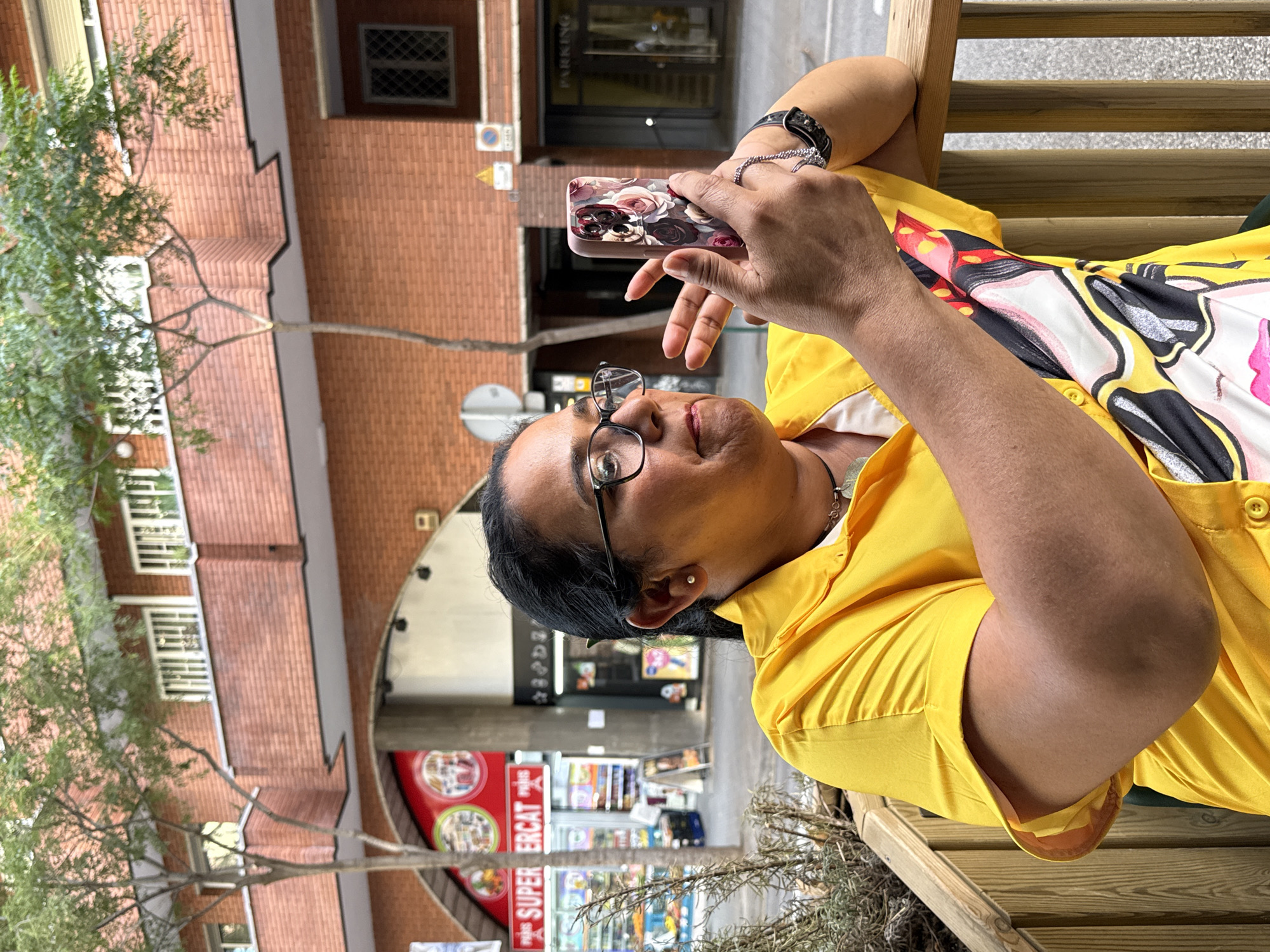 A woman is sitting outdoors at a table, holding a smartphone in her hands. She is wearing glasses, a yellow shirt with a colorful graphic design, and a watch on her left wrist. The background shows a building with brick walls and windows, and a store with a red sign that reads "SUPER ERCAT PARIS". The scene appears to be in a city, possibly Barcelona, Spain, given the "PARIS" text on the store sign which may indicate a chain or a location within Spain.