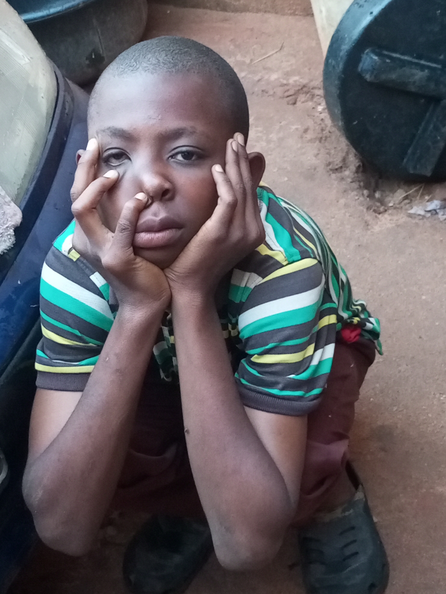 A close-up, eye-level, outdoor, medium shot shows a young boy sitting on reddish-brown dirt with his knees pulled up to his chest. The boy is looking directly at the camera with his hands supporting his face. His eyes are open, and he has a somber expression. He is wearing a t-shirt with horizontal stripes in black, dark green, light green, and yellow. He is also wearing brown pants. His arms are bent at the elbow, with his hands on either side of his face and his thumbs resting on his temples. His fingers are interlaced in front of his face.
The background includes a large, dark blue object on the left, which appears to be the side of a vehicle. Behind the boy, to the right, is a large, black, round object with a textured surface, possibly a container or a barrel. The ground is composed of packed dirt and small pebbles. The lighting suggests it is daytime, with diffused natural light. There are no visible indications of the city of Tempure, Nigeria within the image itself, so its presence cannot be confirmed or denied based on visual evidence alone.