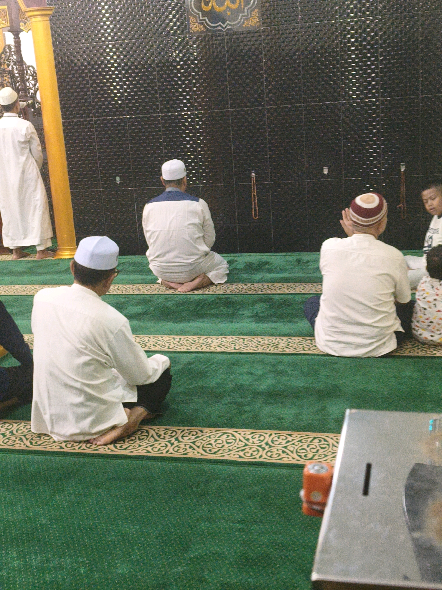 The image depicts the interior of a mosque in Samarinda, Indonesia. Several individuals are engaged in prayer, facing a wall adorned with textured black tiles. A prominent yellow pillar stands to the left of the frame, partially obscuring a figure in a white robe. The floor is covered with green carpeting featuring an ornate pattern along the edges. In the foreground, a metal object with a slot and a circular indentation is visible. Additionally, a small orange object, possibly a battery, rests on the carpet near the metal object. The overall scene suggests a religious gathering and a moment of quiet contemplation.