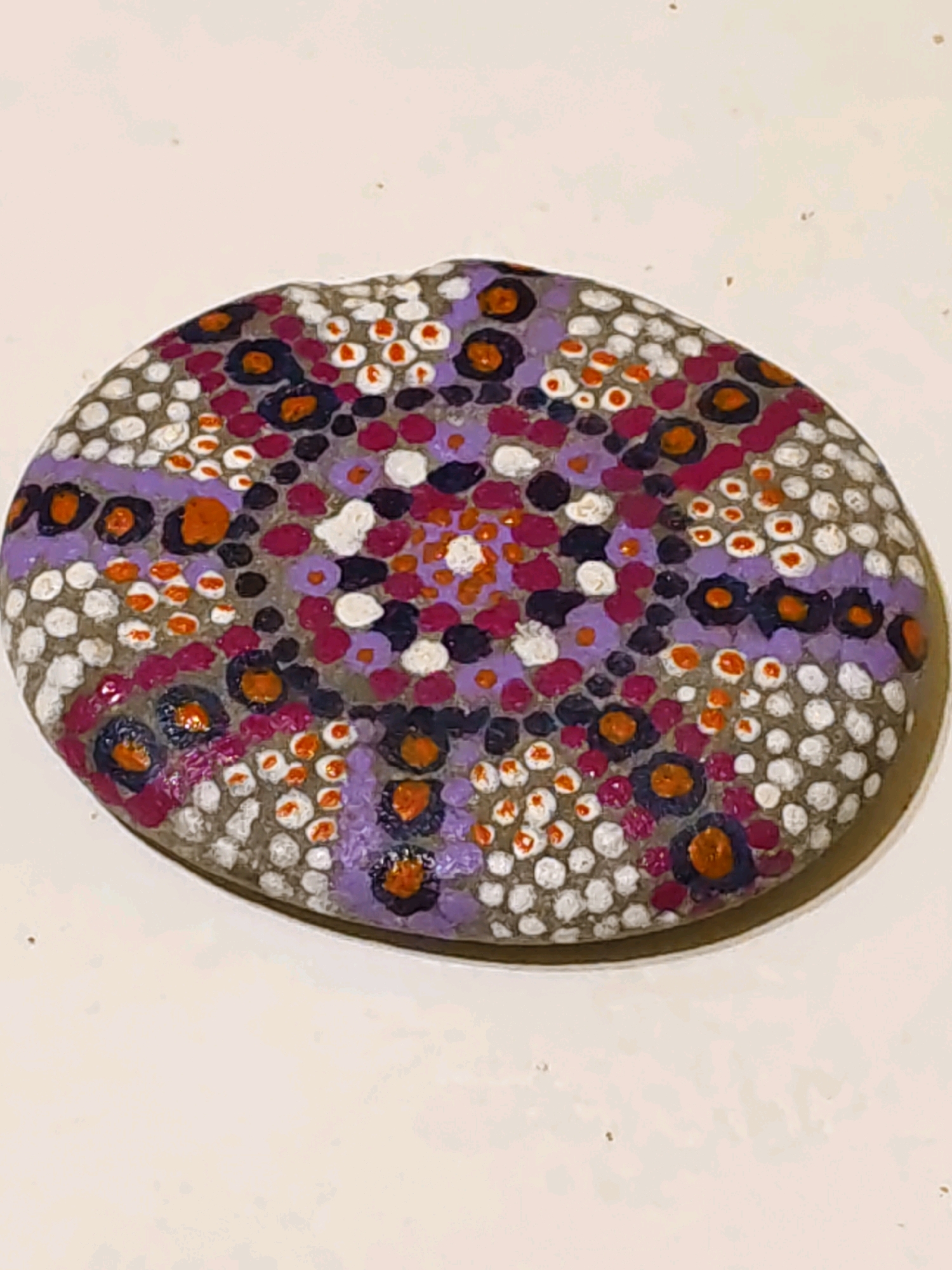The image shows a close-up, top-down view of a flat, oval-shaped rock that has been painted with a dot pattern. The background is a plain, light-colored surface, possibly a table or countertop. The rock itself is gray and appears to be a smooth river stone. The painted design features a variety of small, circular dots in several colors, including white, purple, pink, orange, and dark blue. Some of the dots are arranged in clusters, resembling flowers, while others are scattered more randomly. There are several larger, dark blue dots with orange centers, which stand out against the other colors. The painting style is consistent with pointillism or dot art. The rock is positioned slightly off-center in the frame, with its longer axis running roughly horizontally. The lighting suggests an indoor setting with overhead illumination. There are no people or other objects visible in the image. The image does not contain any information that would allow for its location to be identified as Rostov-on-Don, Russia, or any other specific geographical location.
