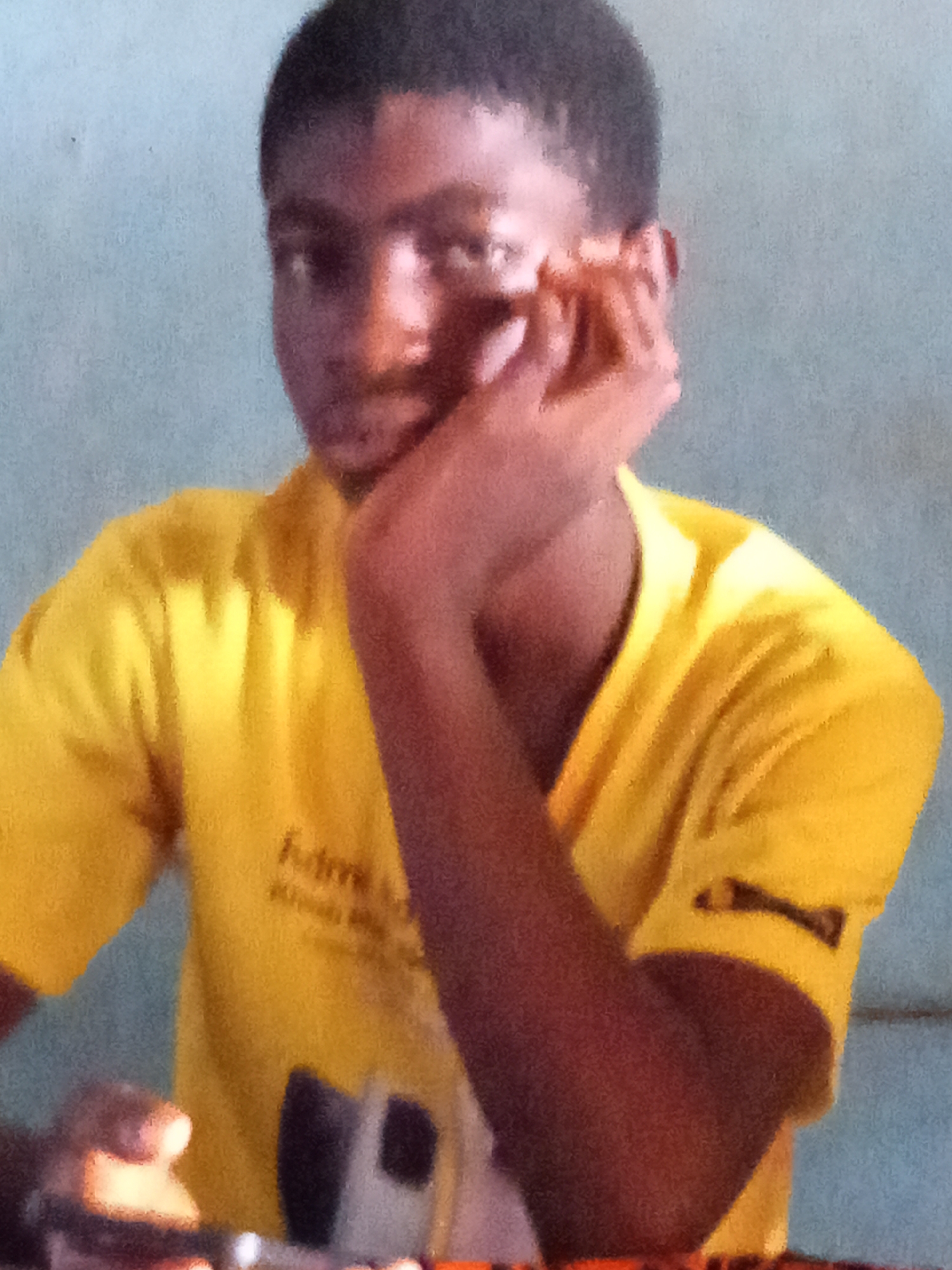 A young man with dark skin and short black hair is resting his face on his hand. He is wearing a yellow t-shirt. The background appears to be a plain wall. The image was taken indoors in what appears to be Nsugpe, Nigeria.