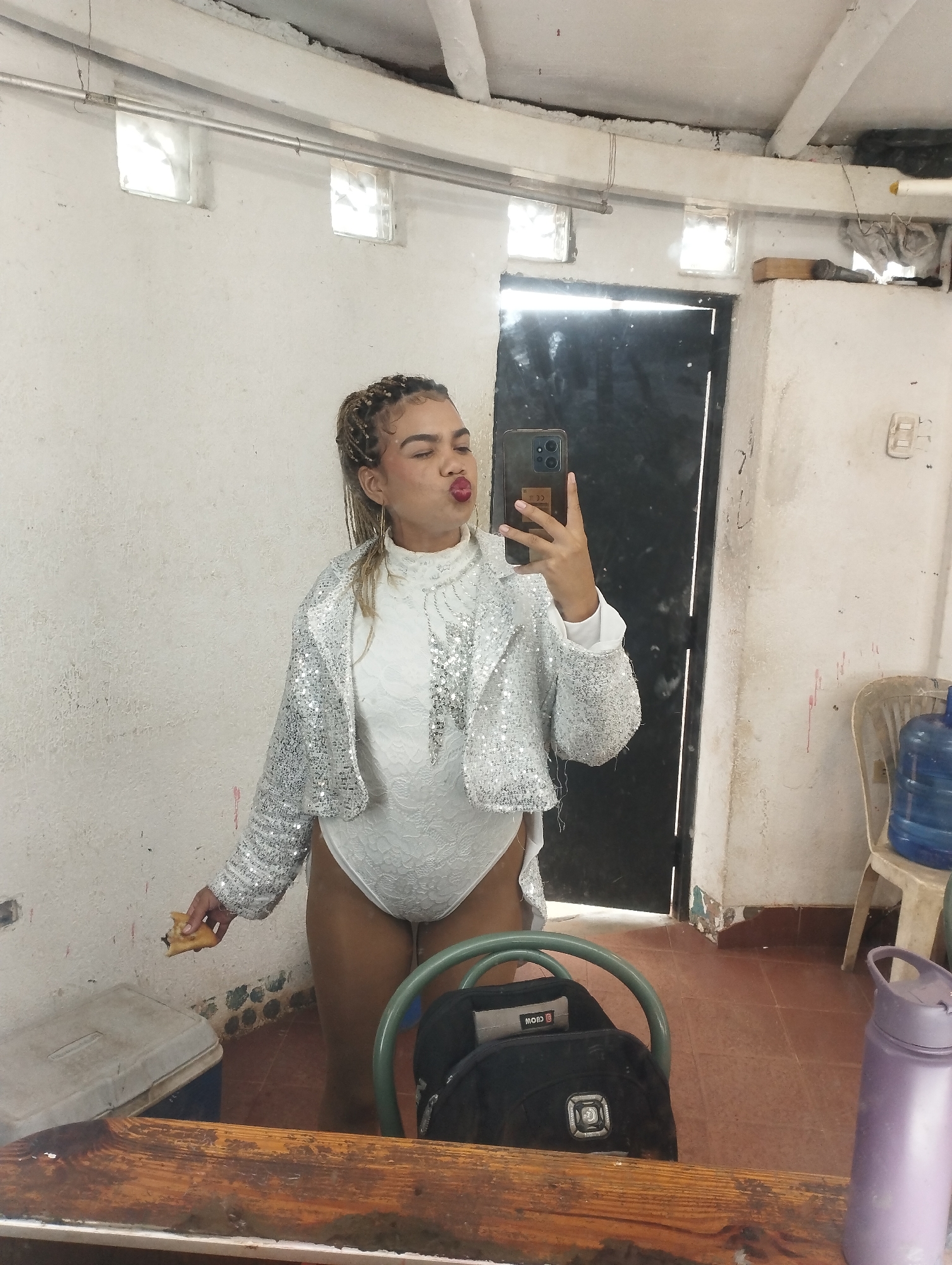 A young woman is captured taking a mirror selfie in an indoor setting. She has braided hair pulled back and is making a playful kissing face towards the camera, holding a dark-colored smartphone in her right hand. She is dressed in a white, high-necked bodysuit with a delicate lace-like pattern, over which she wears a shimmering, cropped silver sequined jacket with long sleeves. In her left hand, she holds a partially eaten pastry or piece of bread with a dark filling.
The background reveals a utilitarian room with light-colored, possibly concrete, walls that show signs of age and wear. A tall, dark door or large mirror reflects a dark space behind her. Near the ceiling, two small glass block windows provide ambient light, and a metal pipe runs horizontally across the upper wall. The floor is tiled in a reddish-brown color.
Various objects are present in the room: a white plastic chair holds a large blue water dispenser jug on the right. An off-white cooler or storage box is visible on the floor to the lower left. In the immediate foreground, a dark backpack with a green metal frame is placed on a wooden surface, likely a table or counter, next to a purple water bottle. A small white label with "MOID" text is visible on the backpack. The overall atmosphere suggests a practical, non-residential space, possibly a dressing room or backstage area.