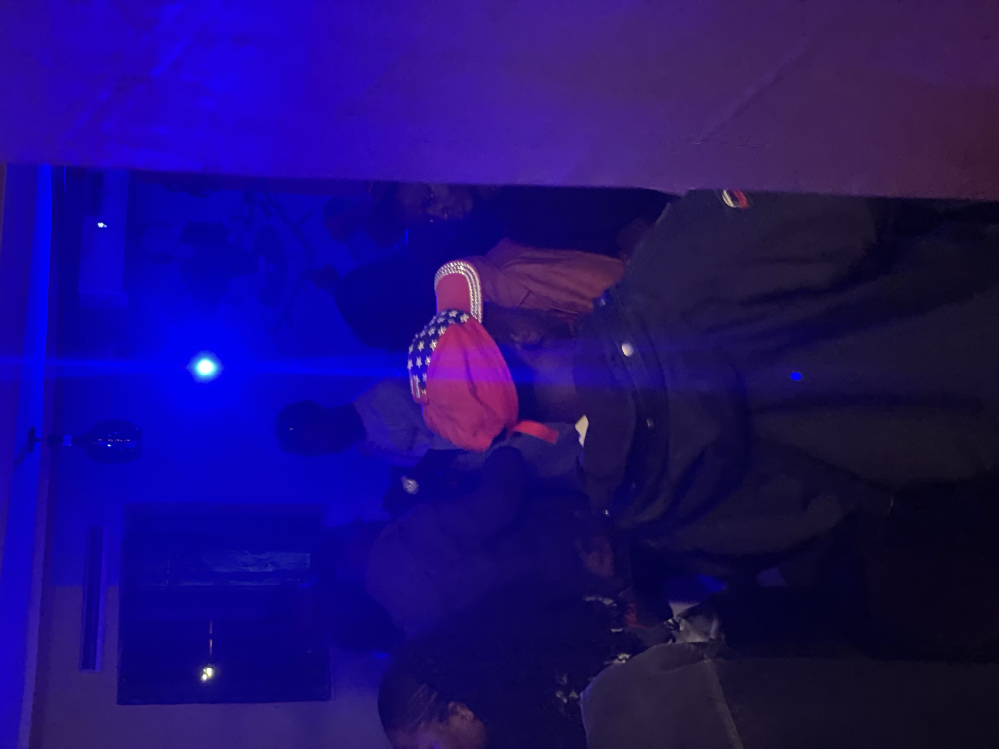 In a dimly lit interior space, possibly a club or social gathering in Durumi, Nigeria, several individuals are present, shrouded in deep blue and purple lighting. A prominent subject in the foreground, facing away from the camera, wears a black jacket and a red hat adorned with a pattern resembling the American flag. The hat has a distinctive sparkly brim. To the left, parts of other individuals are visible, their features obscured by the low light and their positions within the crowd. In the background, faint outlines of more people can be discerned, with one individual appearing to be near a DJ setup. The environment is characterized by the intense artificial lighting, which creates a club-like atmosphere.