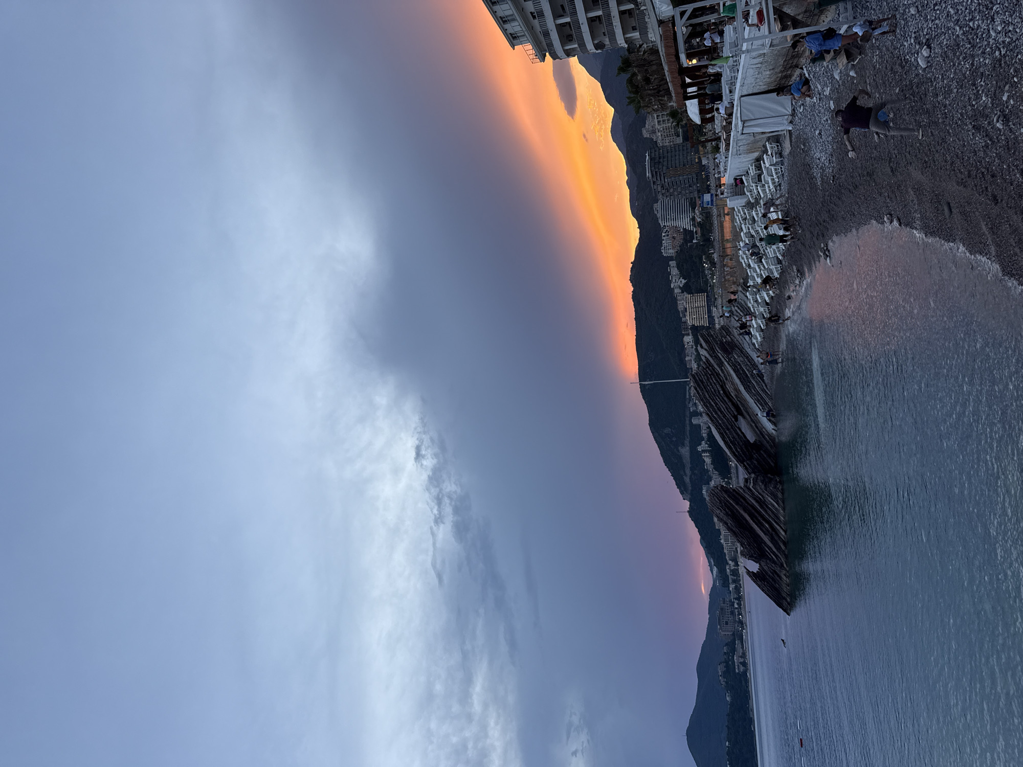 The image captures a scenic view of a beach and coastline at dusk. The sky is filled with dramatic clouds, transitioning from a pale gray on the left to a vibrant band of orange and pink on the right, indicating the sunset. This colorful sky is silhouetted against dark mountains that form the coastline.
Below the mountains, a town or city is visible, with buildings clustered along the shore. A beach occupies the foreground, covered in smooth, dark pebbles. Several white lounge chairs are set up on the beach, some occupied by people. Further out, the calm, dark water of the sea gently laps at the shore.
In the water, a few people are present. One person appears to be swimming or floating. There are also some objects in the water, possibly small boats or buoys. On the beach, a man is lying down on the pebbles. Other individuals are scattered around the beach, some sitting on chairs and others standing. The overall atmosphere suggests a relaxing evening by the sea after a day at the beach. The time of day is clearly sunset, and the weather appears calm with no strong winds.
The image also shows a section of wooden structures jutting out into the water from the beach, adding an interesting element to the coastal landscape. The location is identified as Rafailovići, Montenegro, which aligns with the typical coastal scenery of the region. No visible text is present in the image.