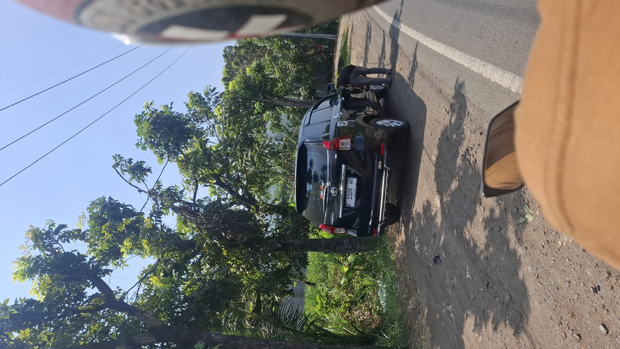 A black Toyota Avanza is parked on the side of a road in Solokpandan, Indonesia. The vehicle has a license plate that reads "F 1108 HR". A person is bent over near the rear passenger side of the car, appearing to be inspecting or working on the tire. The car is situated on a dirt and gravel shoulder next to an asphalt road, with lush green trees and vegetation lining the area. The sky is clear and blue. The image appears to be taken from the perspective of someone on a motorcycle, with a portion of a helmet visible in the foreground on the right side of the frame.