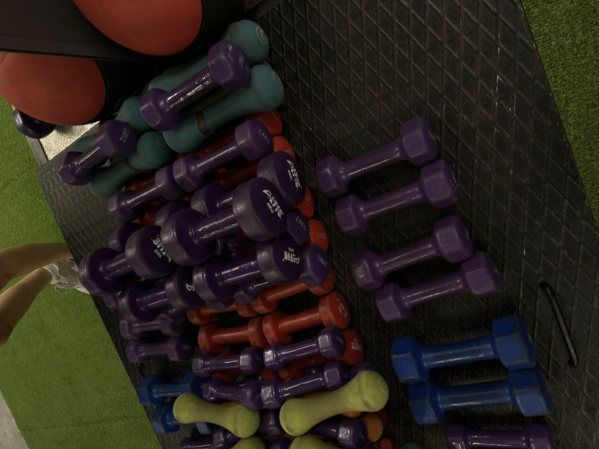 The image presents a close-up, angled view of dumbbell racks in a gym setting. The racks are filled with dumbbells of varying weights, identified by labels like "2.0KG" and "4KG." The dumbbells come in different colors, including purple, teal, red, blue, and yellow-green. The brand name "ALTIS" is visible on some of the dumbbells. The background consists of gym flooring, transitioning from a black textured surface to green artificial turf. Part of a person's leg and shoe are visible on the left side of the image, close to the green turf. The setting appears to be indoors within a fitness facility, but there is no location of Yerevan, Armenia mentioned.