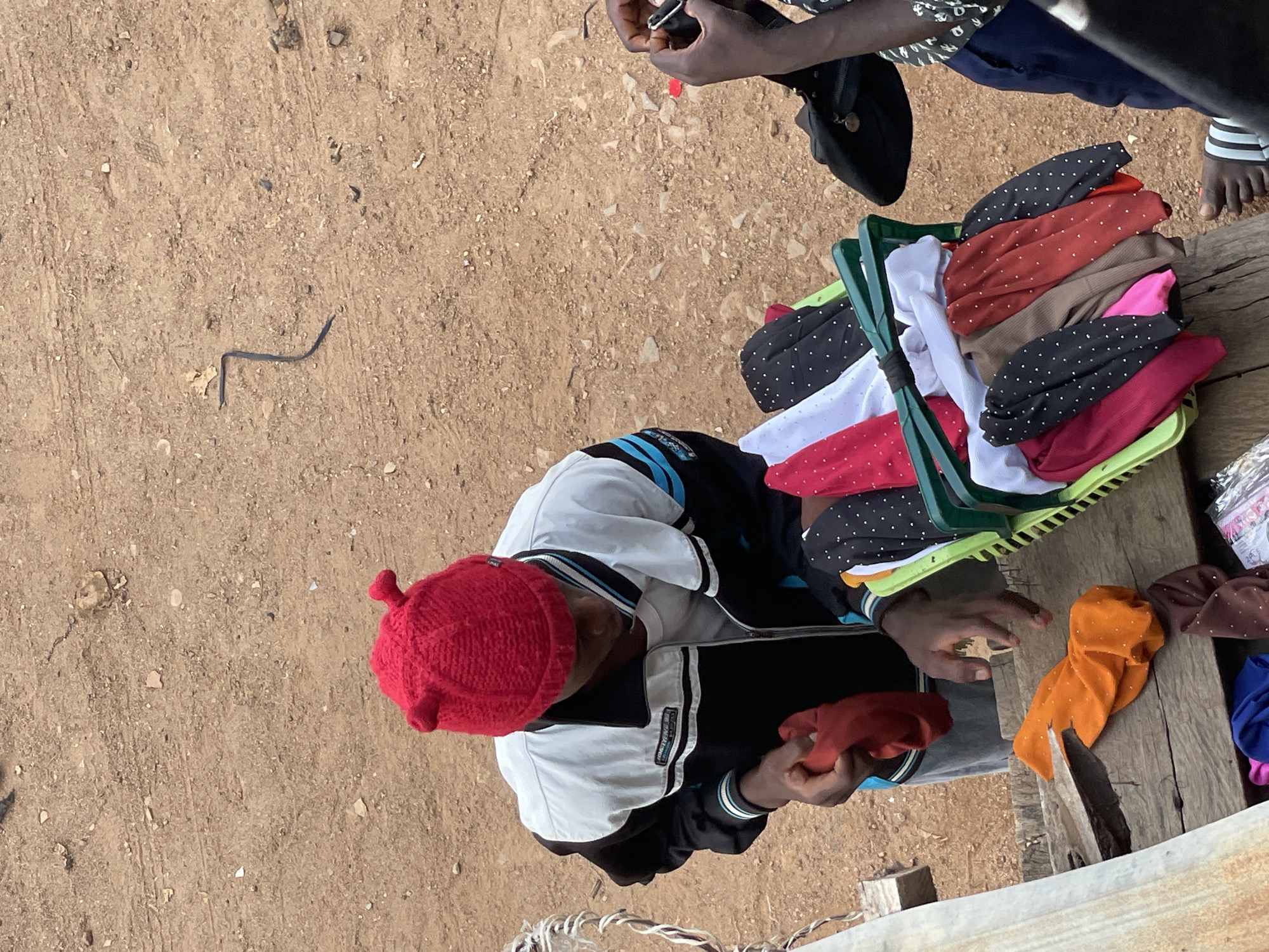 The image depicts an outdoor scene, possibly in an urban area of Unknown, Unknown. The ground appears to be dirt. In the foreground, a person wearing a red knit cap and a jacket is seated, handling pieces of fabric or clothing. A green plastic basket filled with folded items, including polka-dotted textiles in various colors, is present. Adjacent to the basket, more clothing items are arranged on a wooden surface. Another person, partially visible, is interacting with the first person.