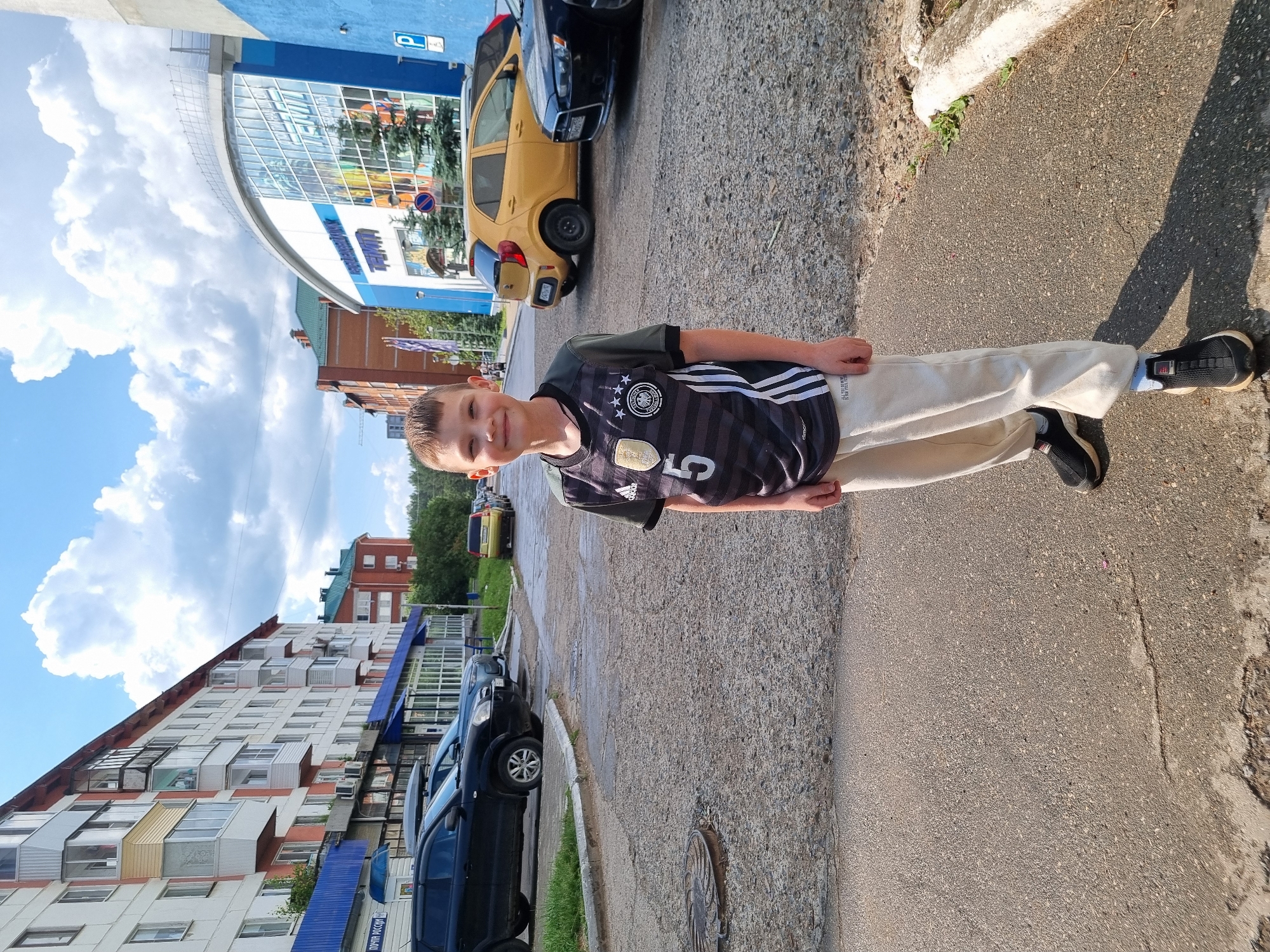 A young boy wearing a black and grey soccer jersey with the number 5 and light-colored sweatpants smiles at the camera. He is standing on a cracked asphalt road in front of apartment buildings and a modern building with a blue and white facade. Several cars are parked on the side of the road, including a black SUV, a yellow hatchback, and a black sedan. The sky is blue with fluffy white clouds. The scene appears to be a residential area in the city of Chaikovsky, Russia.