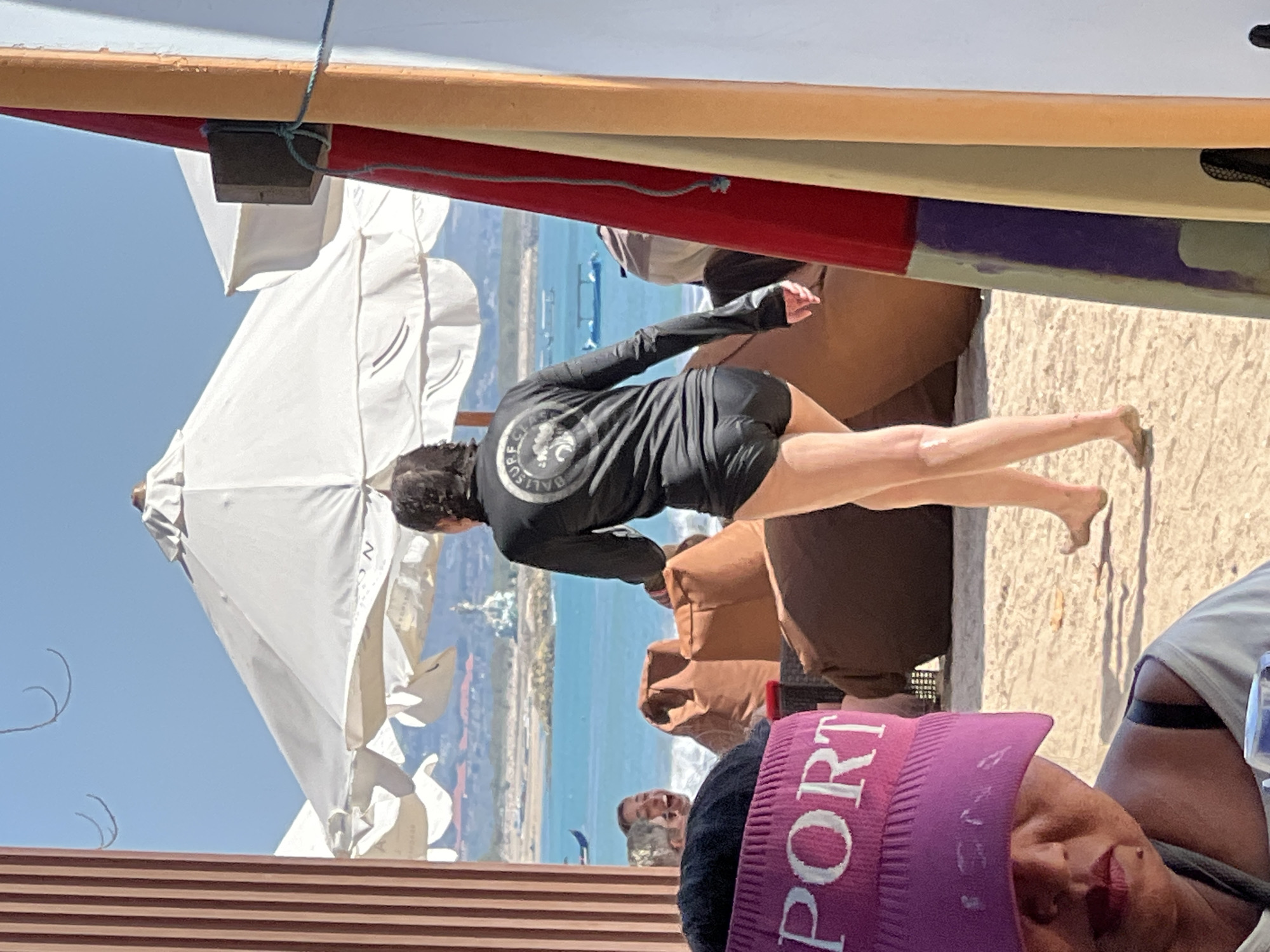 A person is walking away from the camera on a beach in Kuta, Indonesia, wearing a black long-sleeved rash guard with a circular logo and black bottoms. The person's legs are visible, and they are walking towards the ocean. In the foreground, a woman wearing a pink visor with the word "PORT" in white letters is visible, looking down. Behind the woman, there are brown beanbag chairs and white umbrellas. The ocean is blue with some boats in the distance. The scene appears to be set in a beachside cafe or resort.